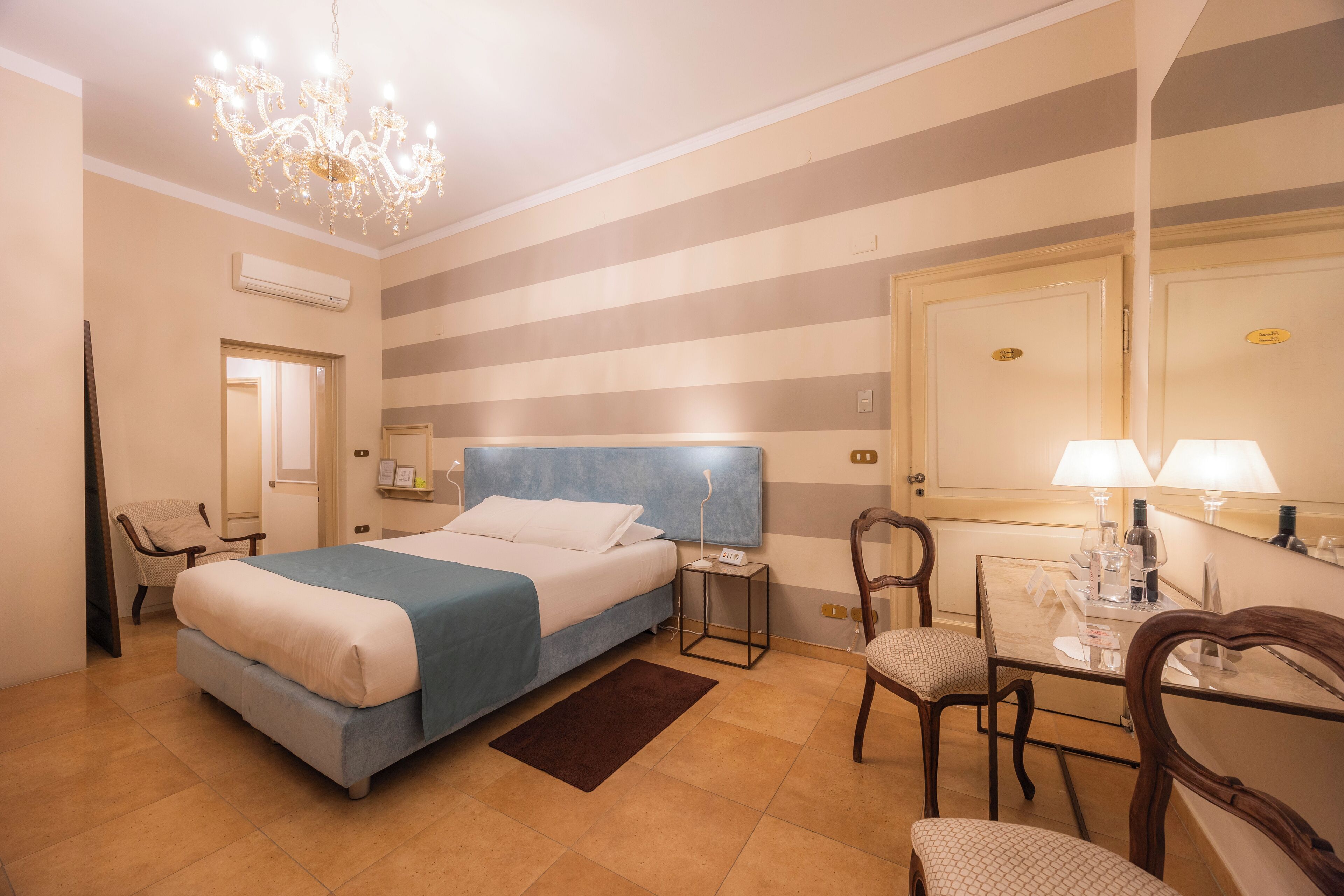 A hotel room with a bed, bedside table, chairs, a desk, a chandelier, and a mirror.