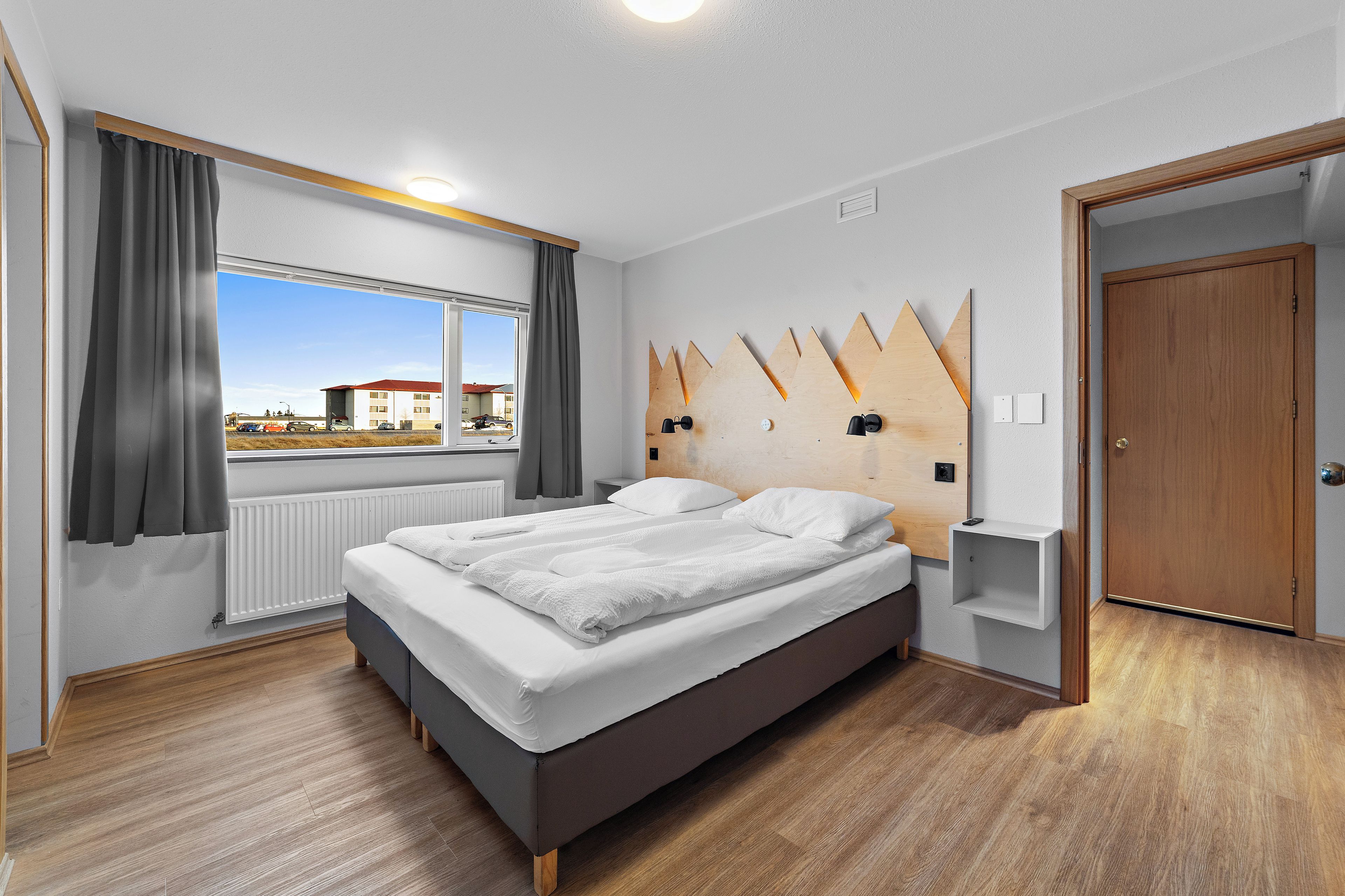 Habitación de hotel moderna con una cama grande, ventana con cortinas, cabecero de madera y vistas al exterior.