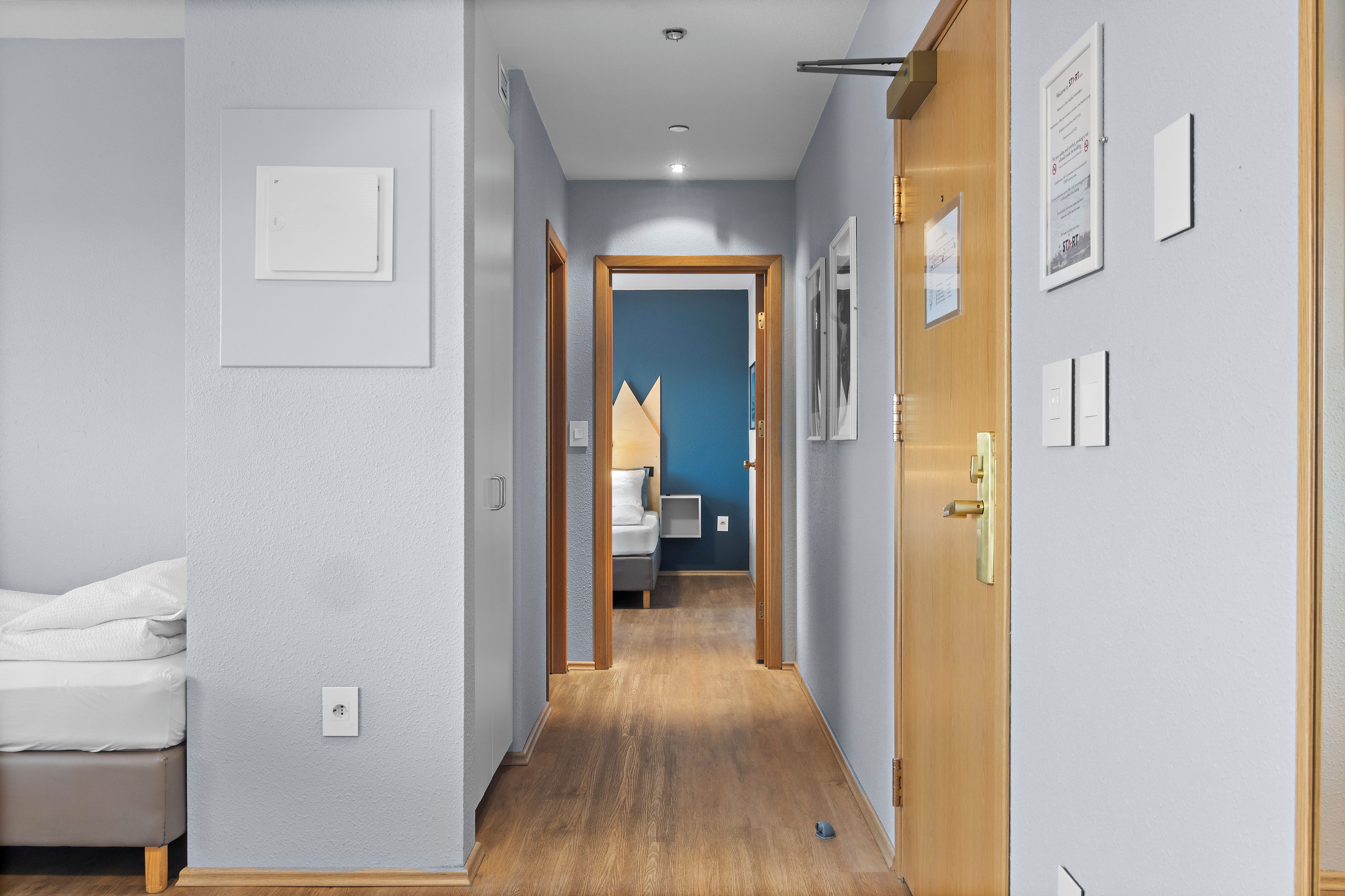 Um corredor com uma cama à esquerda, uma porta à direita e uma parede azul ao fundo.
