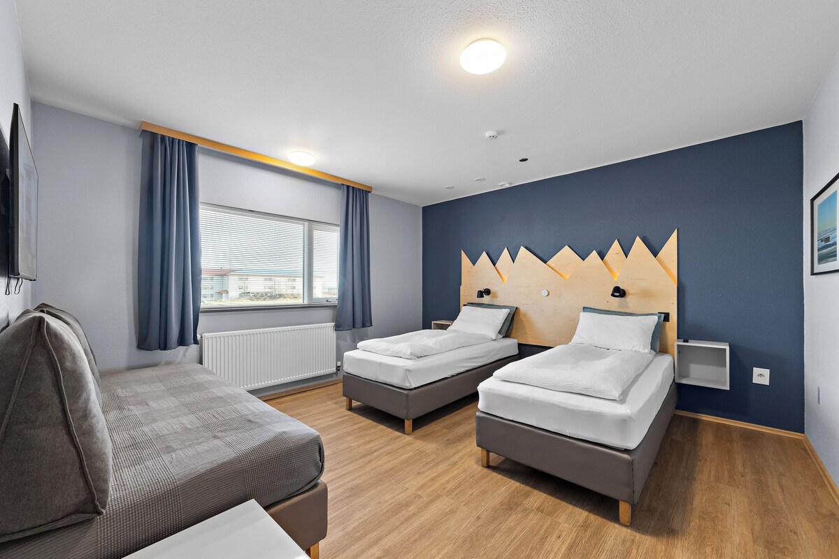 A hotel room with two single beds, a sofa, a window with curtains, a radiator, and a wall-mounted light fixture.