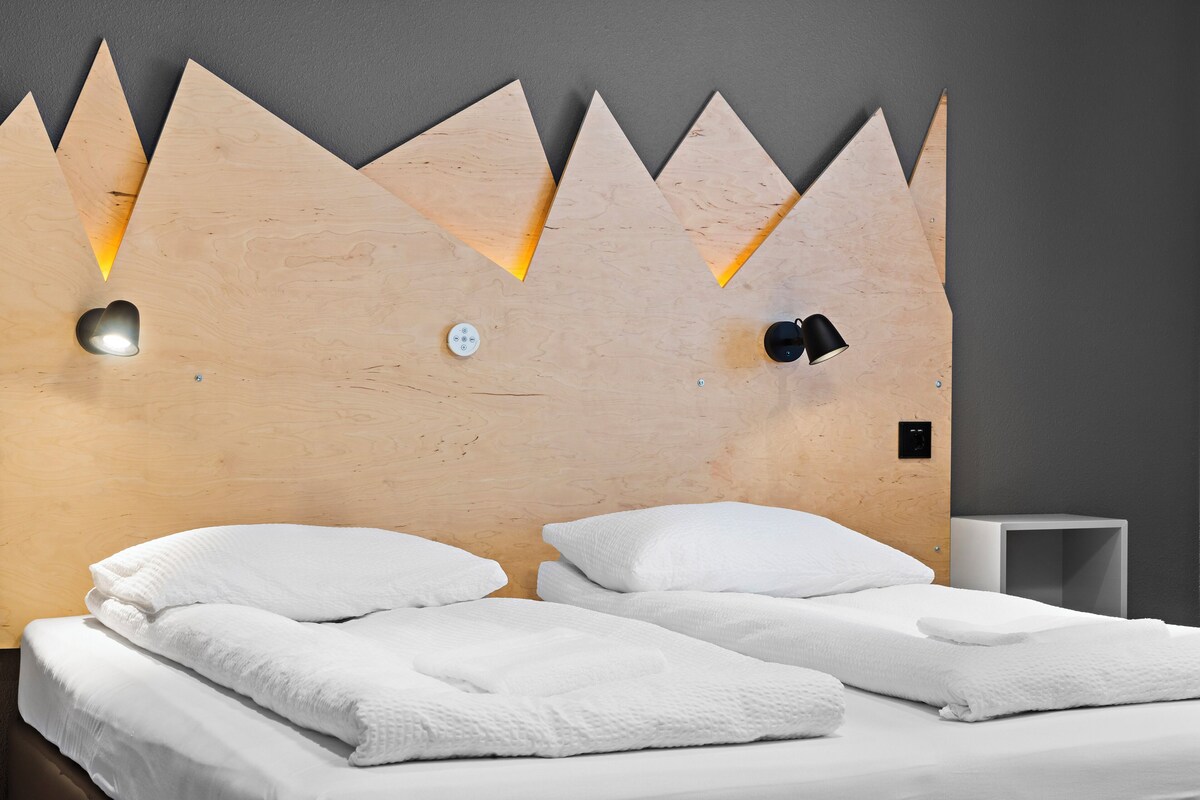 A modern hotel room with a unique wooden headboard, two beds with white linens, and a small bedside table.