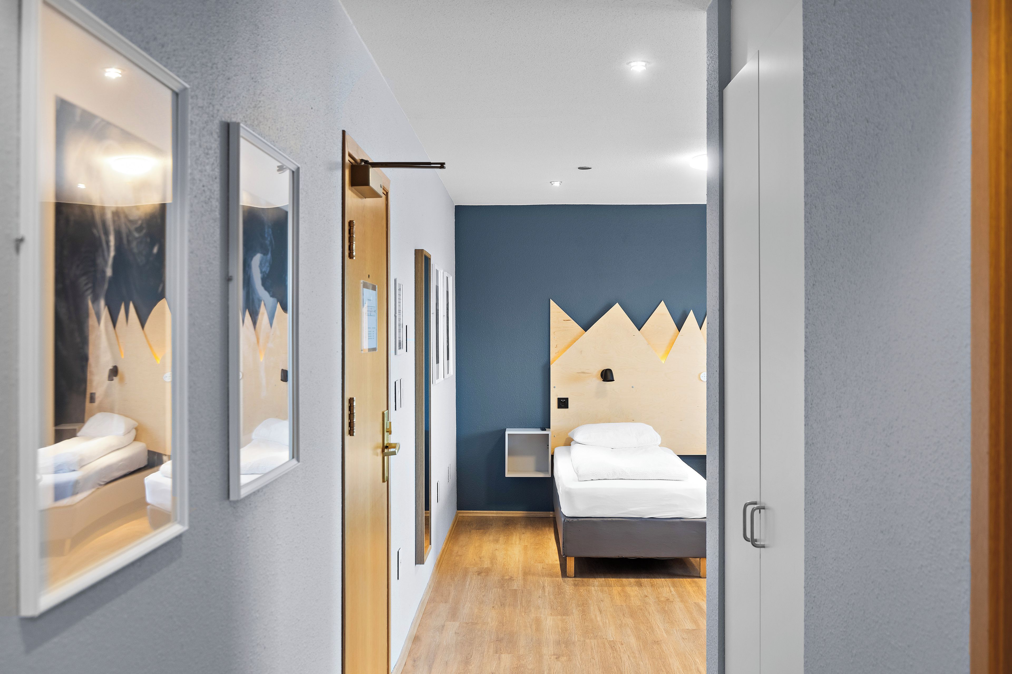 Habitación de hotel con cama, mesita de noche, lámpara de pared y puerta que da a otra habitación.