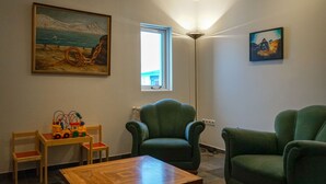 A room with a wooden table, two green armchairs, a painting of a seascape, and a window with a view of a building.