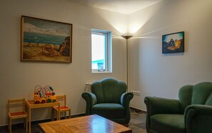 Une pièce avec une table en bois, deux fauteuils verts, un tableau représentant un paysage marin et une fenêtre donnant sur un immeuble.