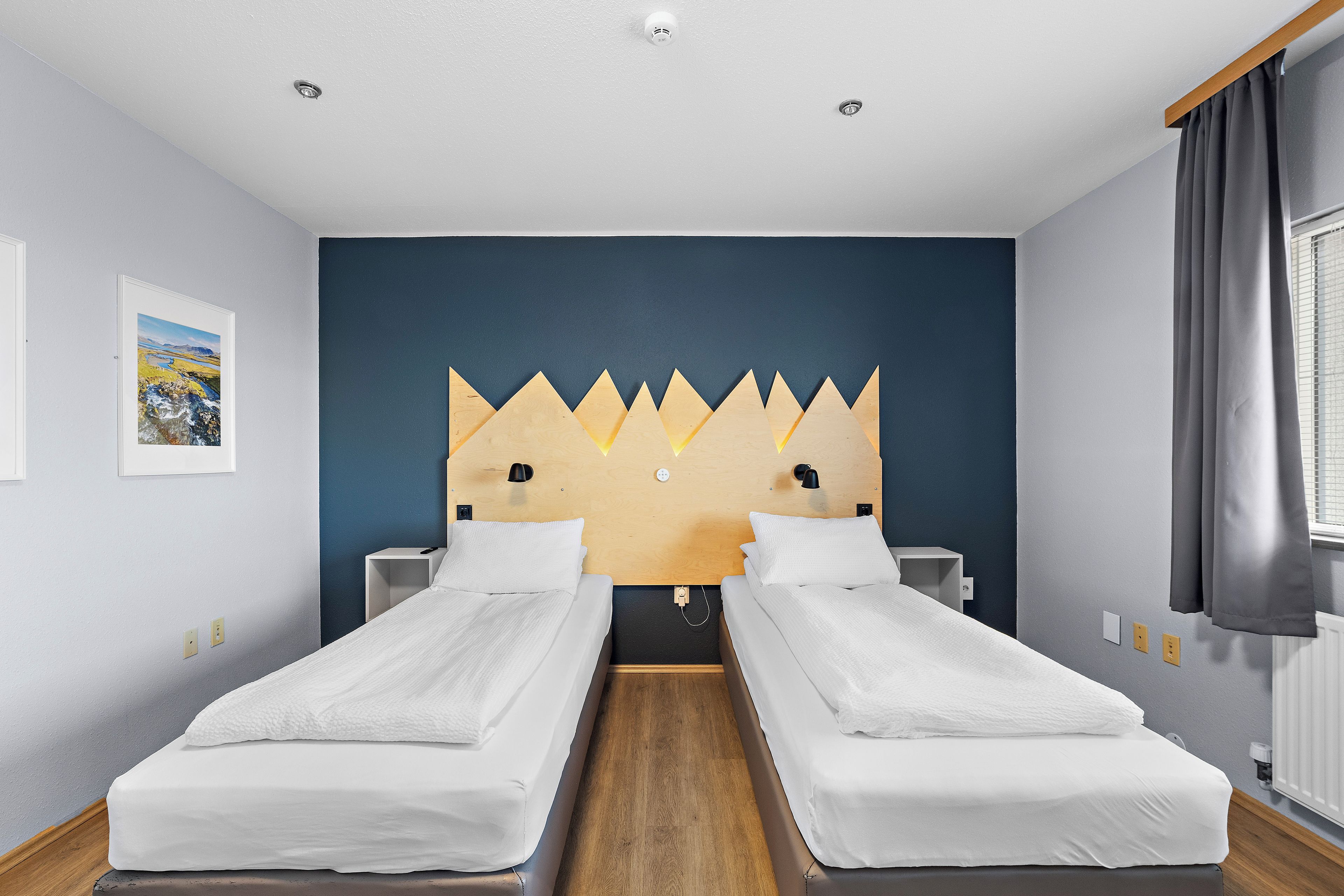 A hotel room with two single beds, a wooden headboard, a painting on the wall, and a window with curtains.