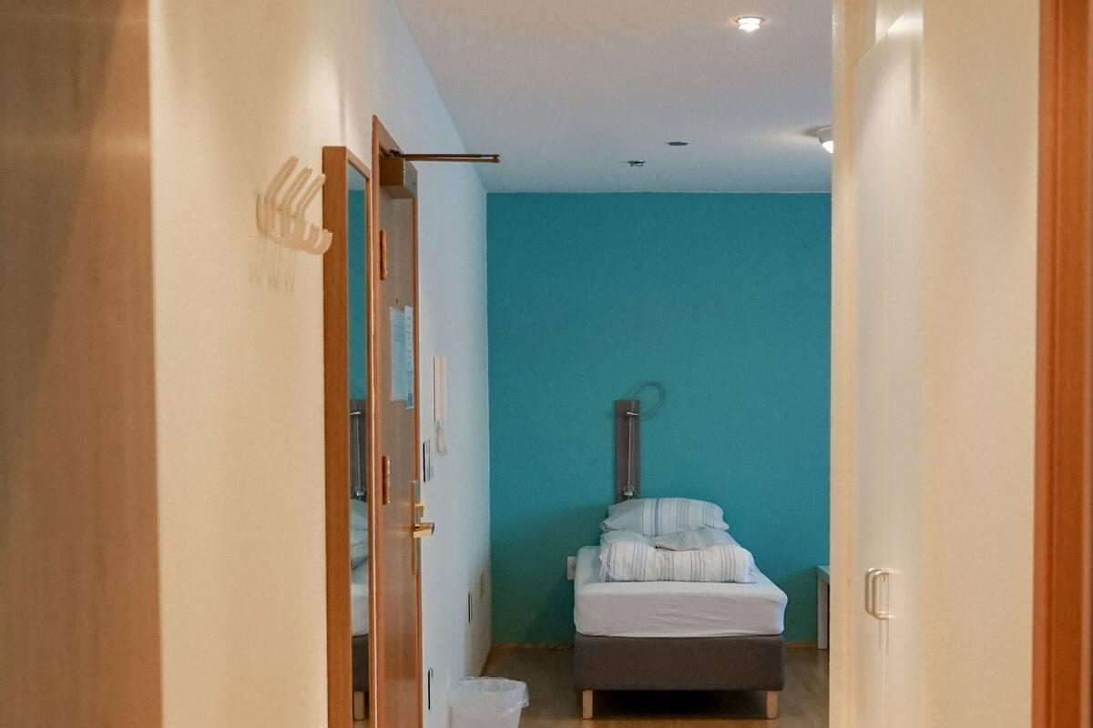 A narrow hallway with wooden flooring leading to a bedroom with a bed and a blue wall.