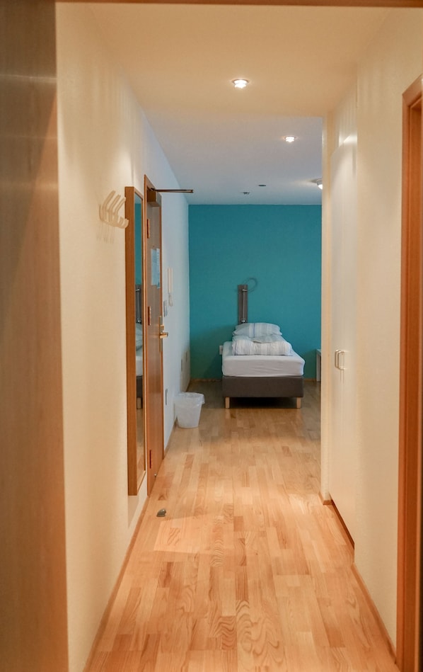 Un couloir étroit, doté d’un plancher en bois, mène à une chambre à coucher où se trouvent un lit et un mur bleu.