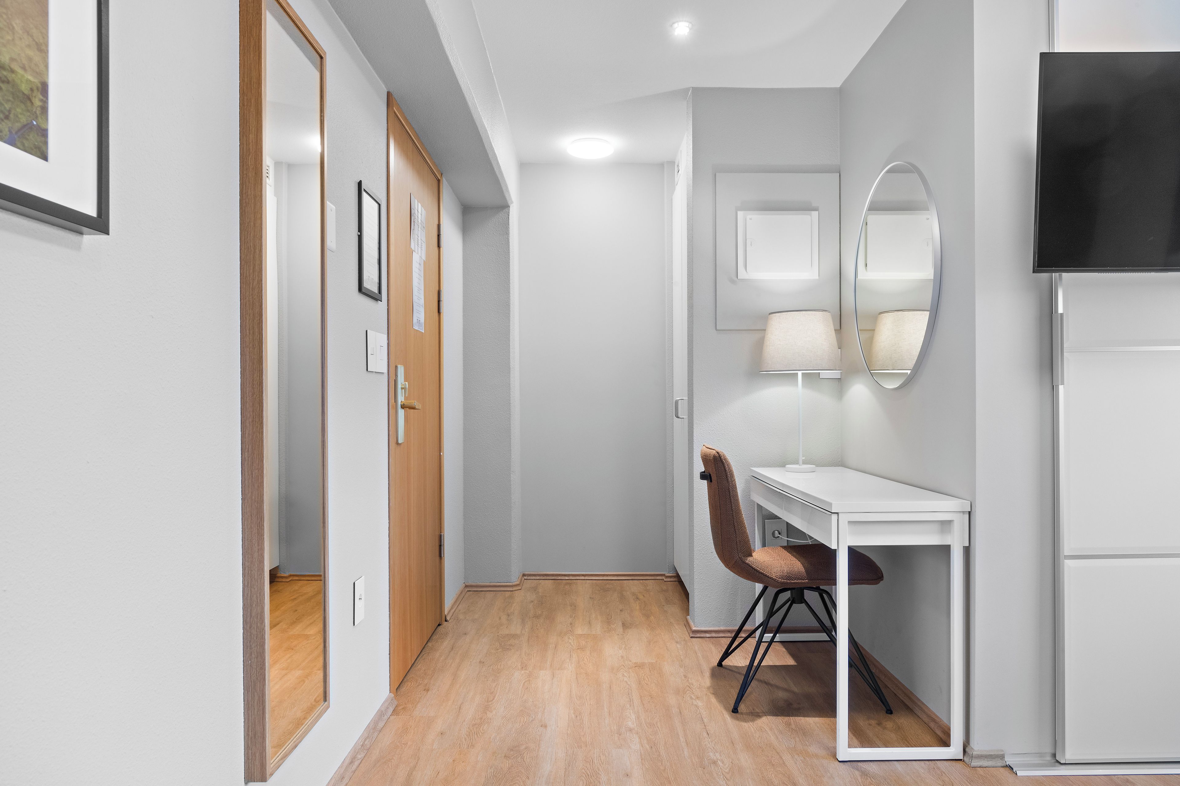 A modern hotel room with a wooden floor, a desk with a chair, a flat-screen TV, and a mirror on the wall.