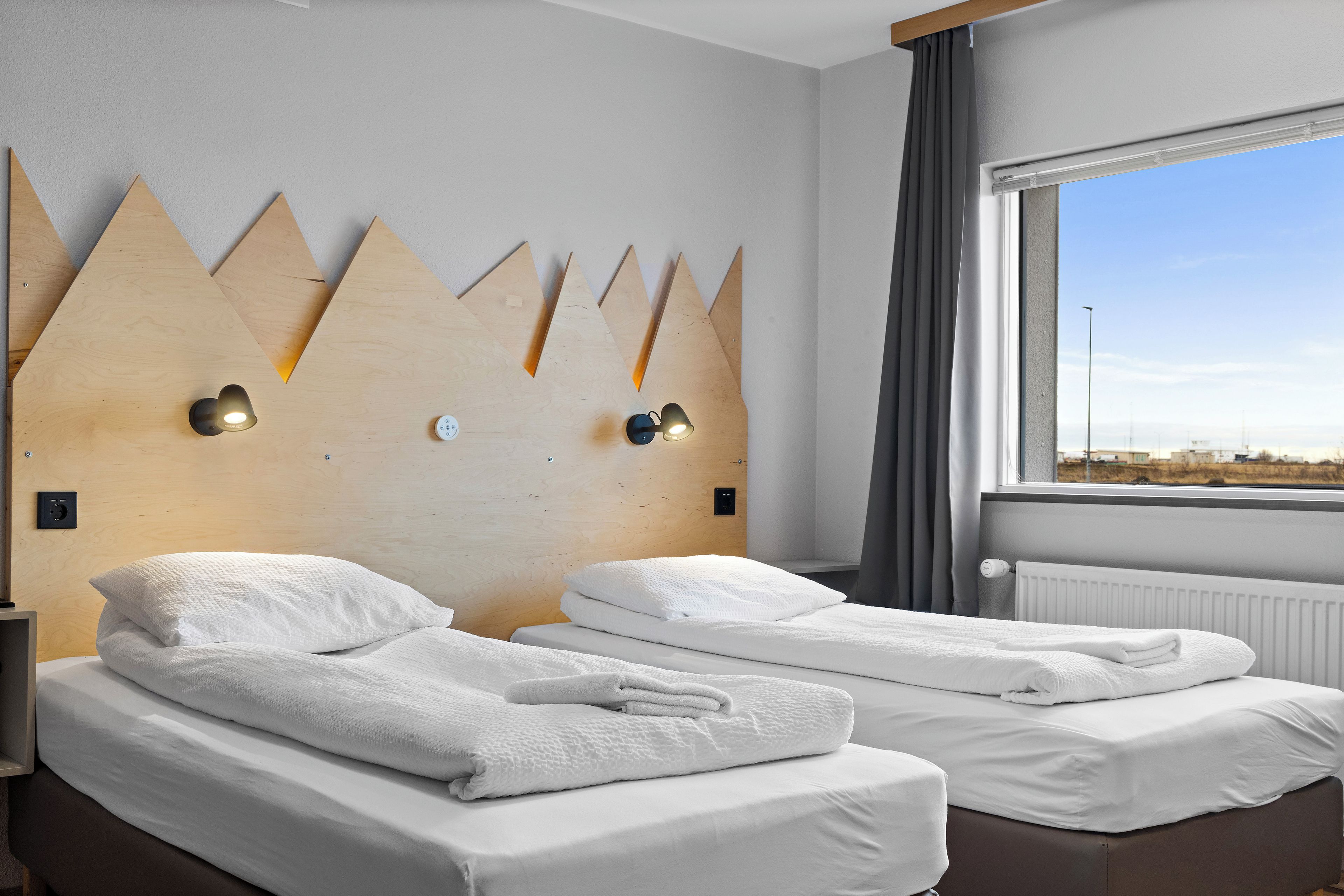 A hotel room with two beds, a wooden headboard with light fixtures, a window with a view, and a radiator.