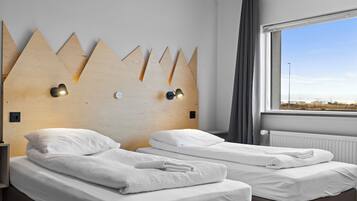 A hotel room with two beds, a wooden headboard with light fixtures, a window with a view, and a radiator.