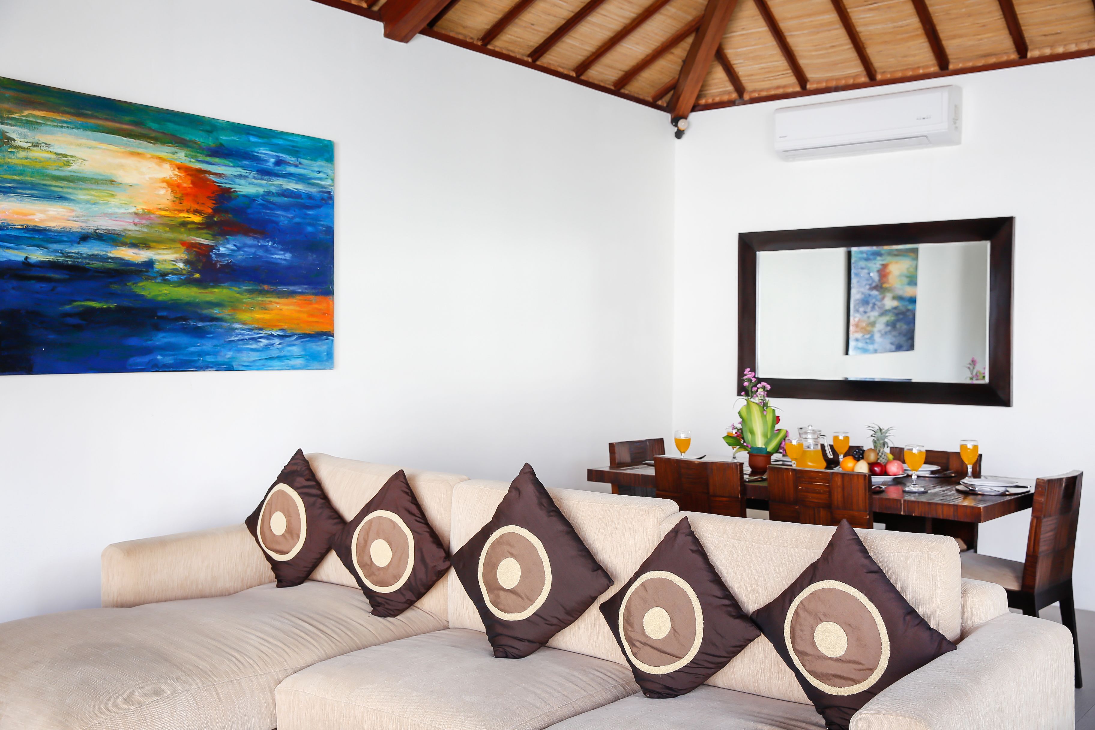 Una sala de estar con un sofá color beige, almohadones decorativos, un cuadro, un comedor con una mesa dispuesta para dos personas y un espejo grande en la pared.