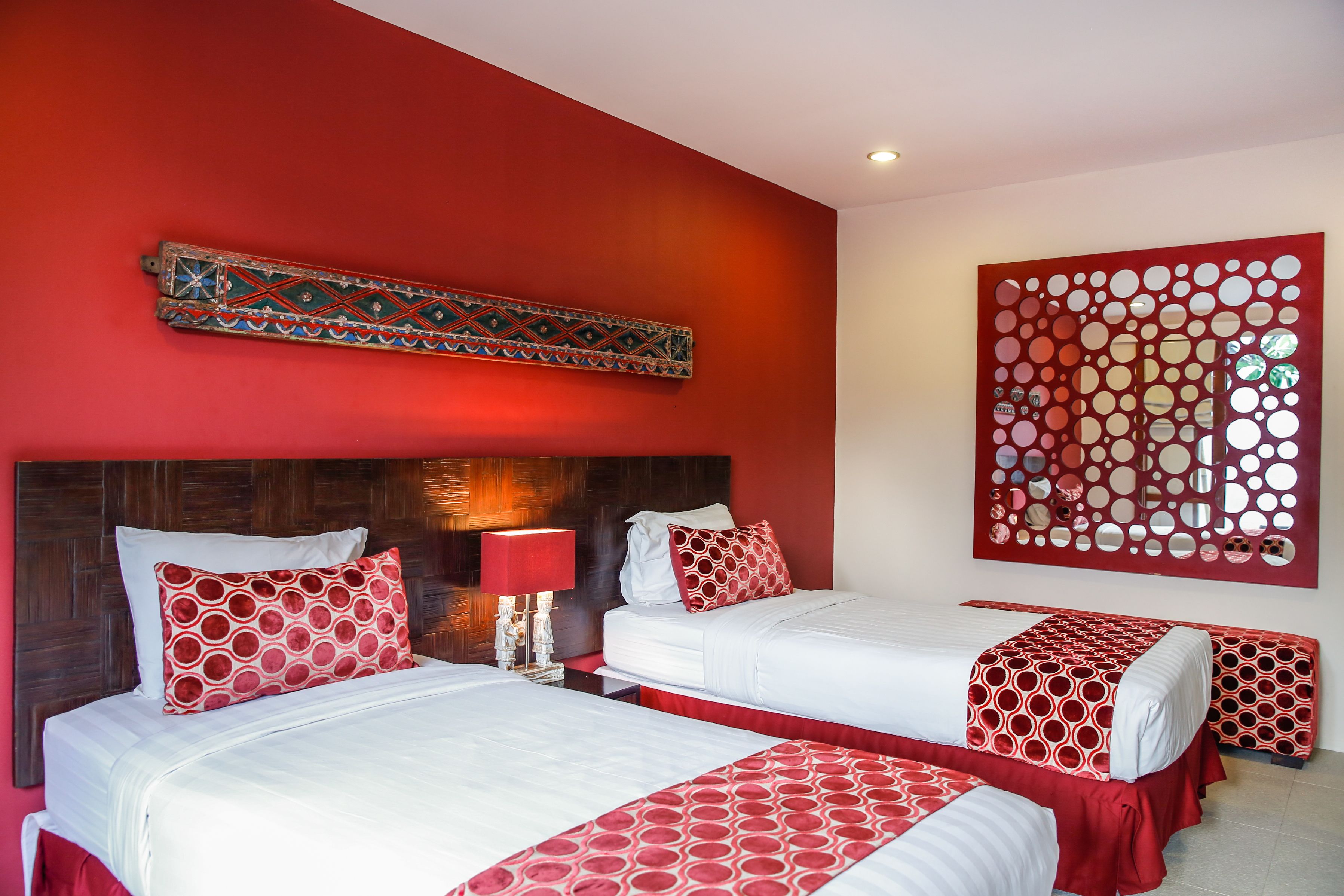 A hotel room with two beds, red and white bedding, a decorative headboard, a wall-mounted mirror, and a red decorative panel.