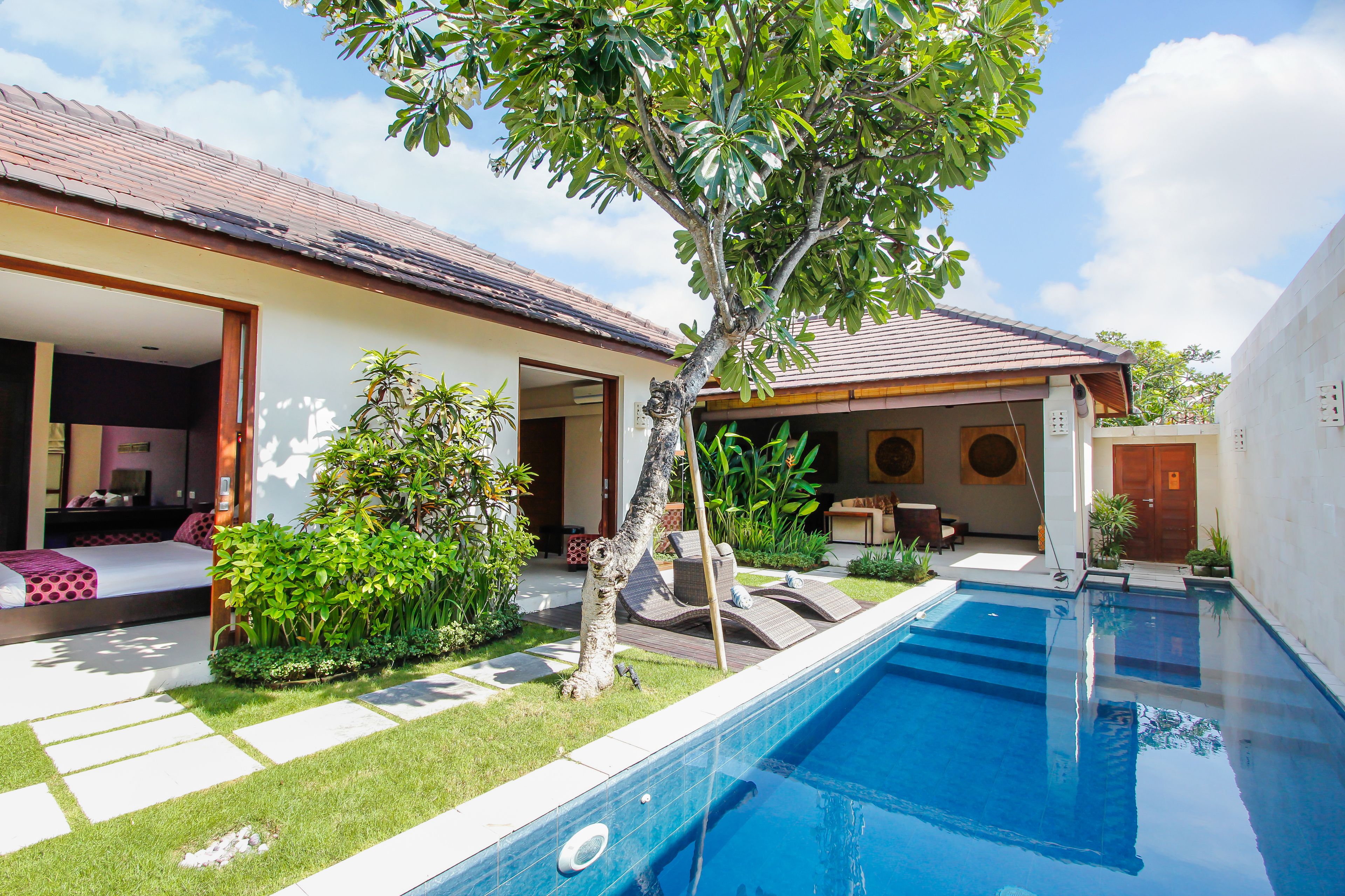 A swimming pool with a tiled deck, a small garden area with a tree, and a building with an open door.