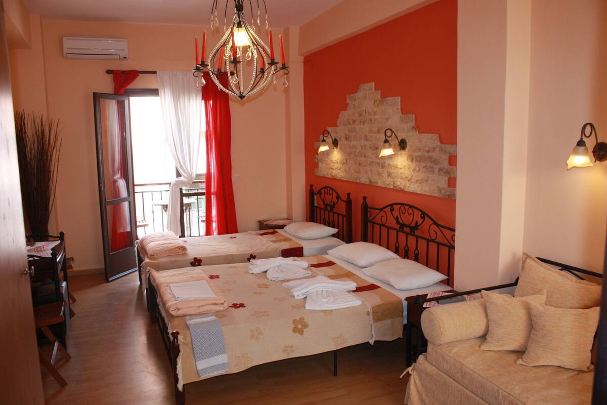 Une chambre d’hôtel avec deux lits, un lustre, un mur d’accent rouge et un balcon avec un rideau rouge.