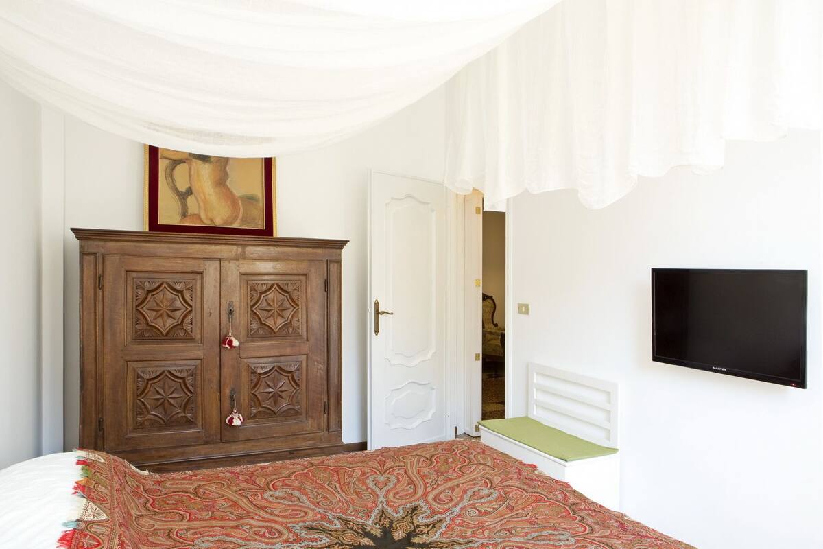 Un dormitorio con un amplio armario de madera, una cama con colcha estampada, un televisor en la pared y una puerta que da a otra habitación.
