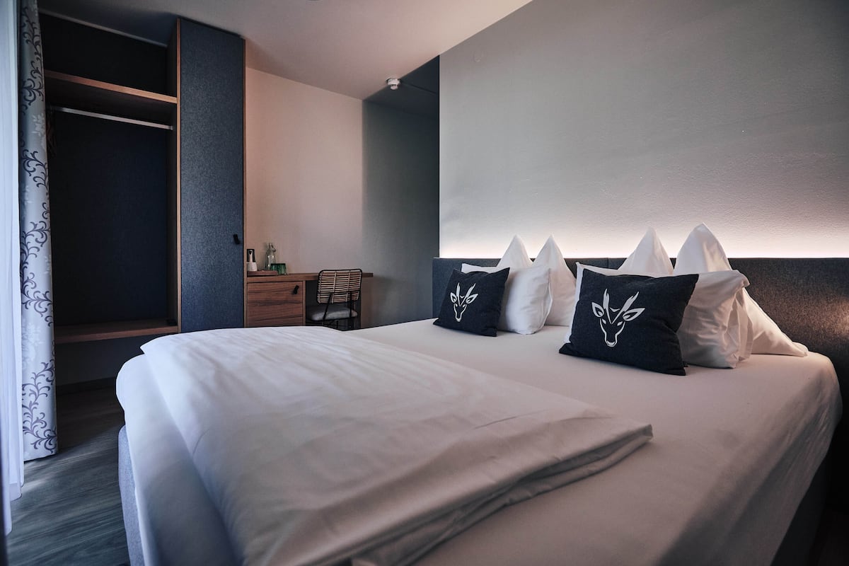 Una cama bien hecha con ropa blanca y almohadones decorativos, una mesita de noche de madera con lámpara, y un armario con espejo.