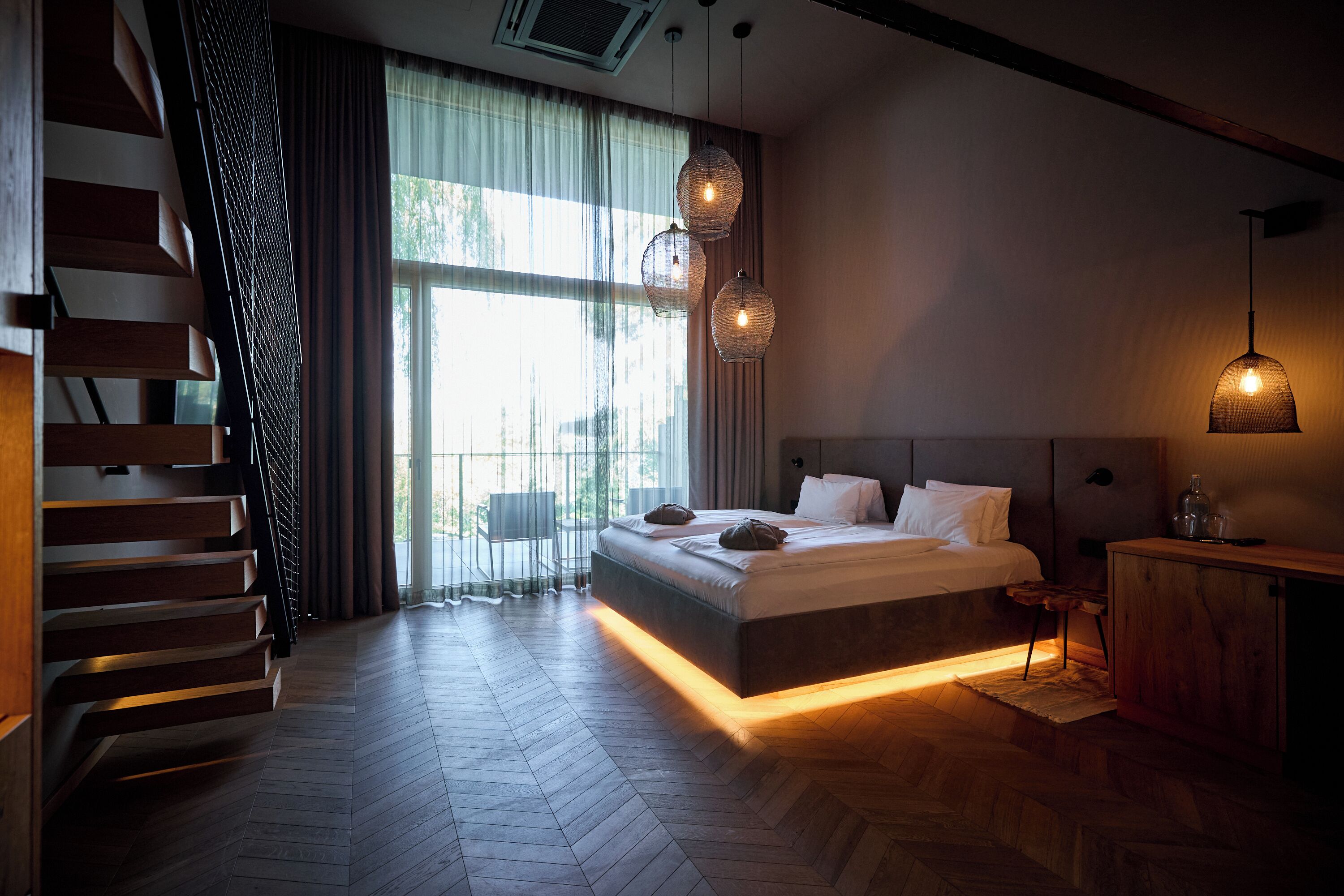 A modern bedroom with a large bed, a wooden staircase, and a view of the outdoors.