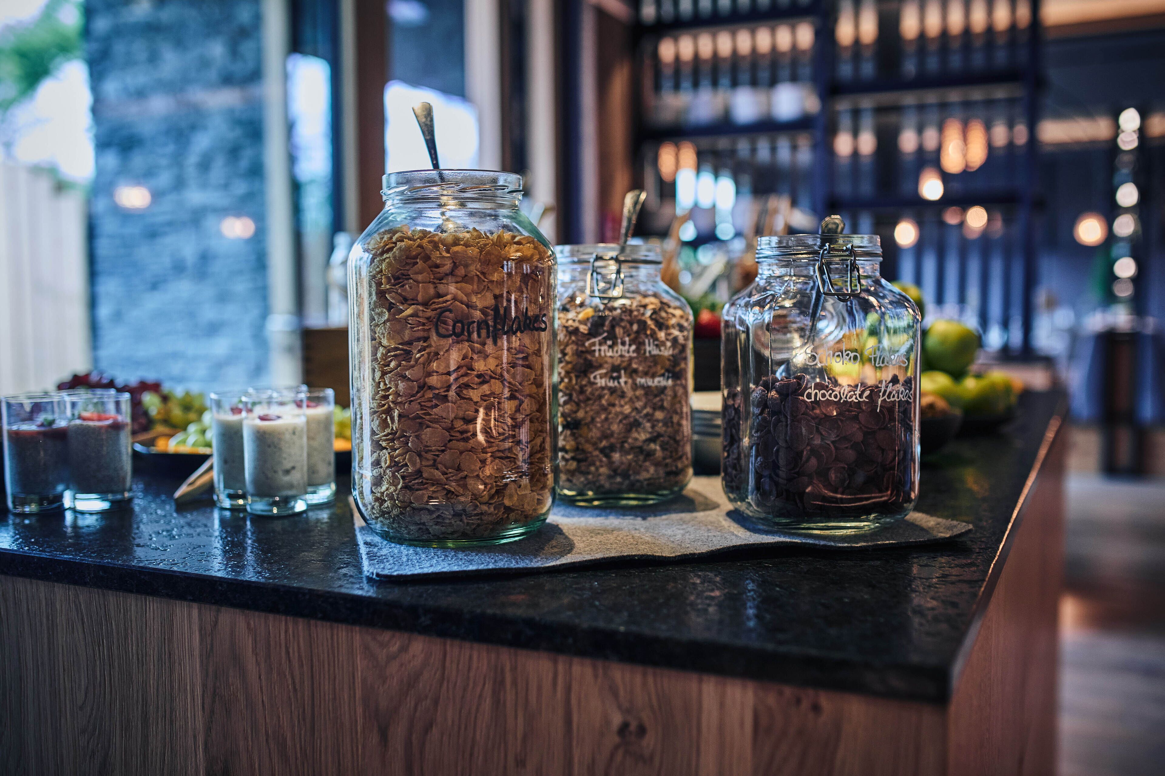 Una barra con varios recipientes de aperitivos y bebidas, como copos de maíz, mezcla de frutas y trozos de chocolate.