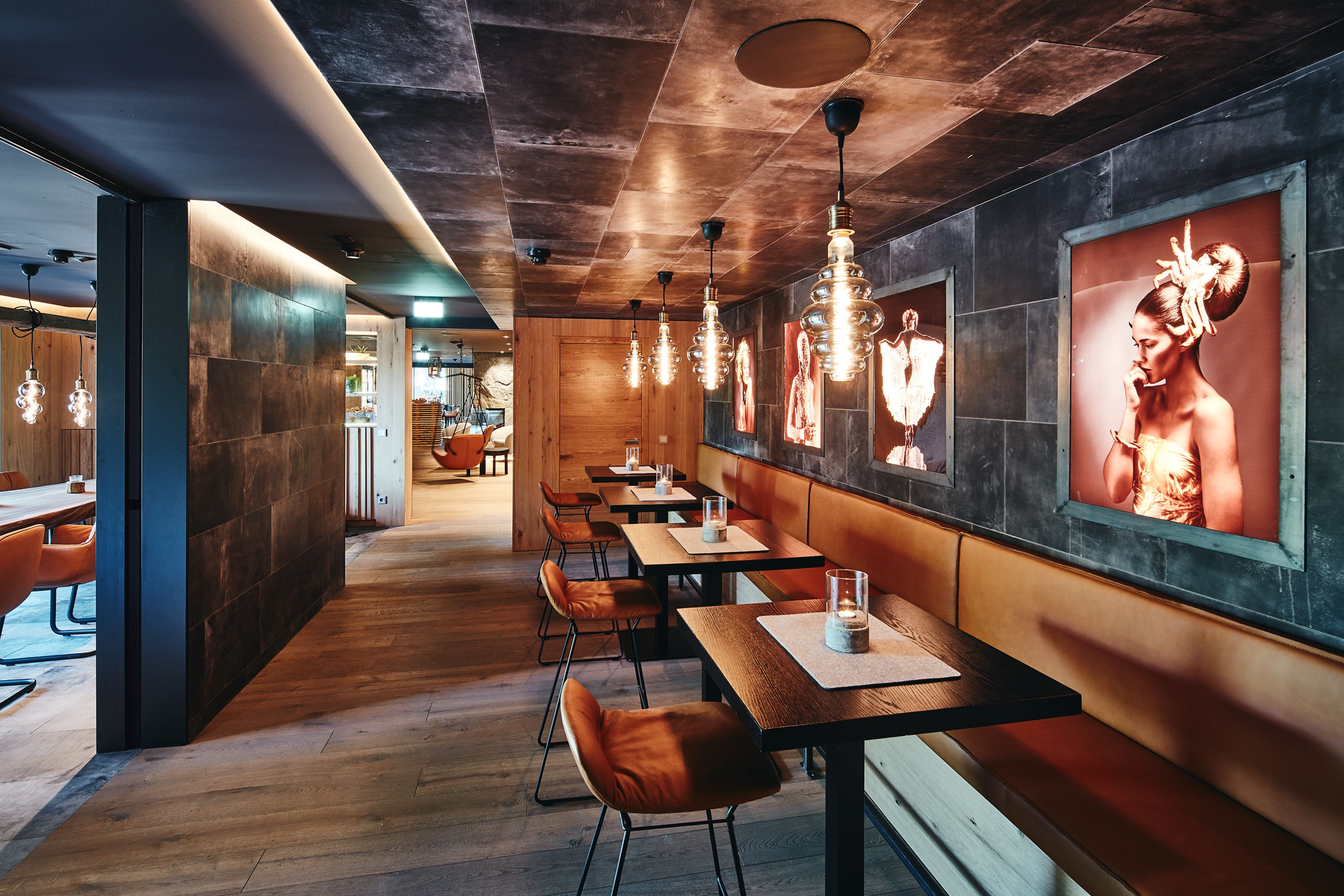 A modern restaurant interior with wooden tables, leather chairs, hanging pendant lights, and large framed artwork on the walls.