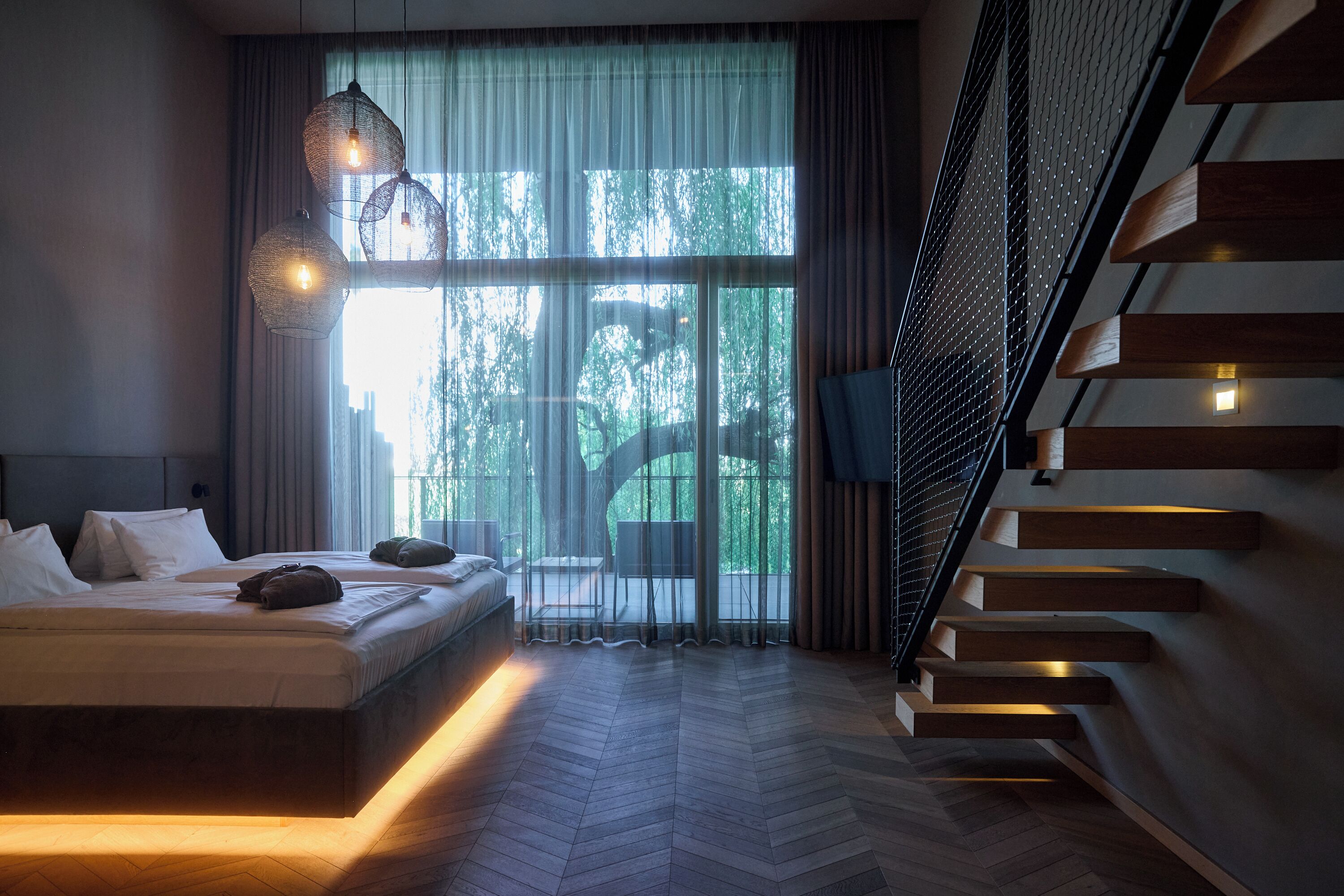 A modern bedroom with a large bed, a staircase, and a view of greenery outside.