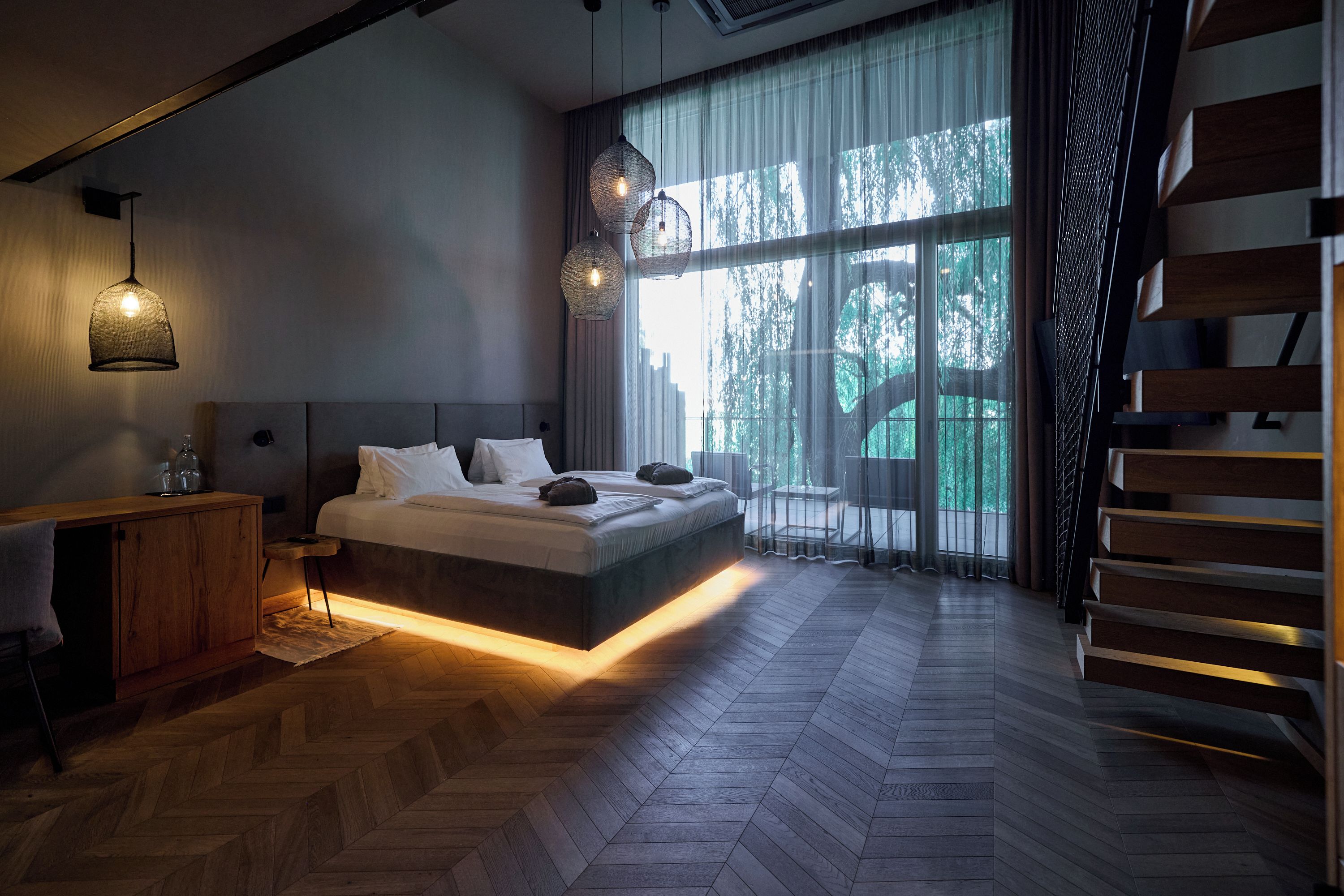 Ein modernes Schlafzimmer mit einem großen Bett, Holzboden und Blick auf Bäume durch ein großes Fenster.