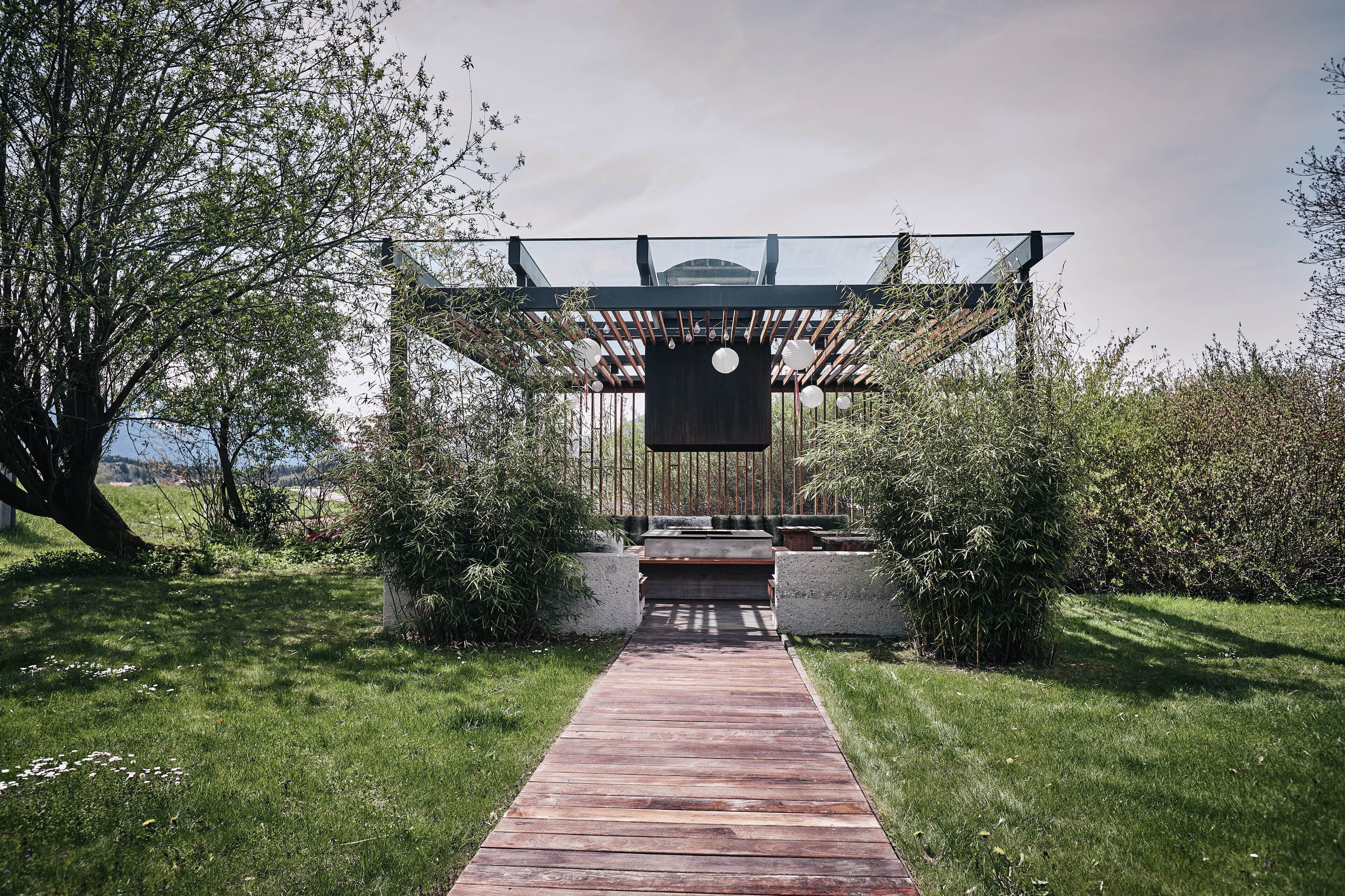 Un sendero de madera que conduce a una moderna pérgola con techo de vidrio, rodeada de exuberante follaje y un cielo despejado.