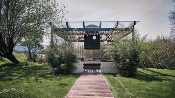Un sendero de madera que conduce a una moderna pérgola con techo de vidrio, rodeada de exuberante follaje y un cielo despejado.