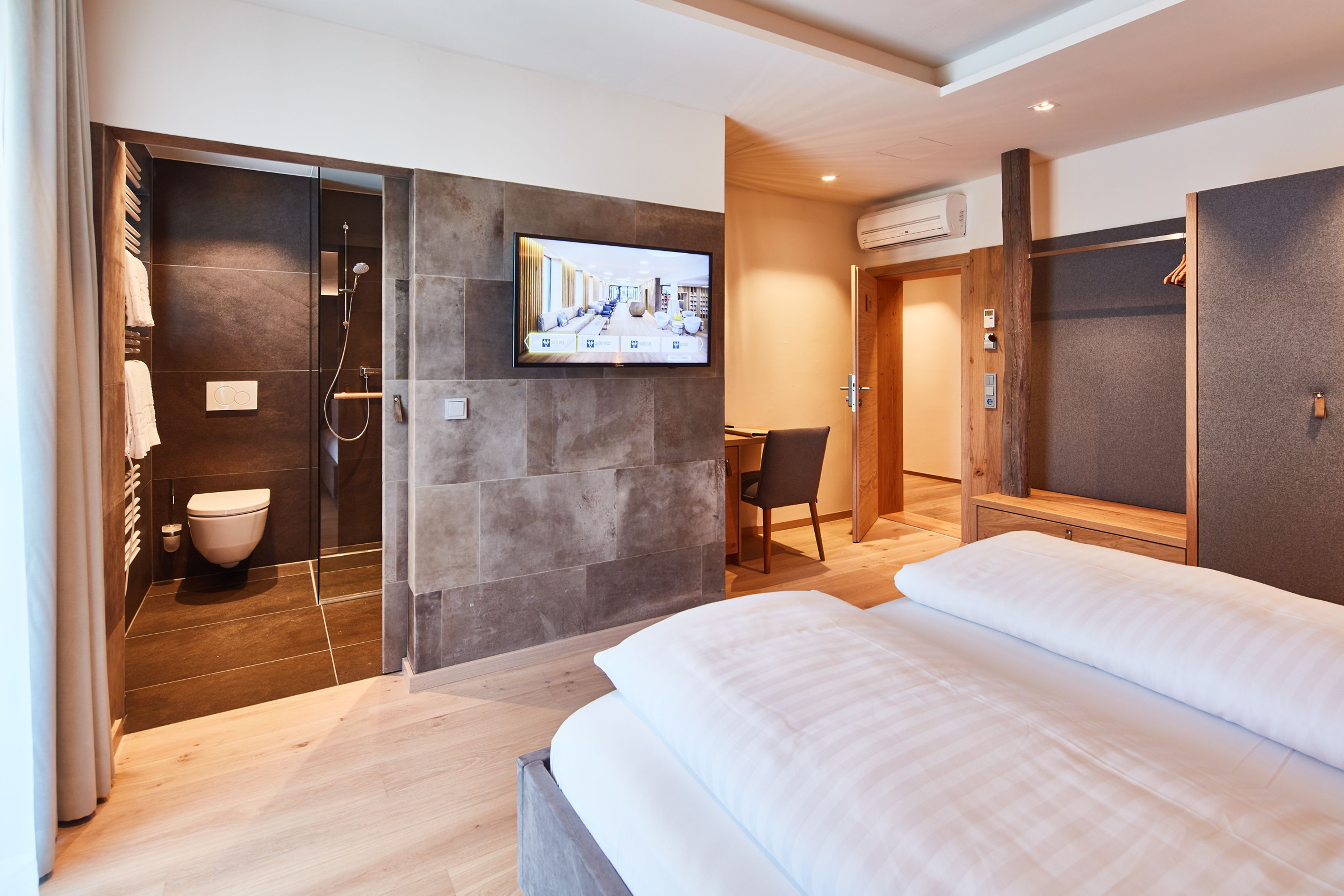 A modern hotel room with a large bed, a TV mounted on the wall, a shower area, and a wooden desk.