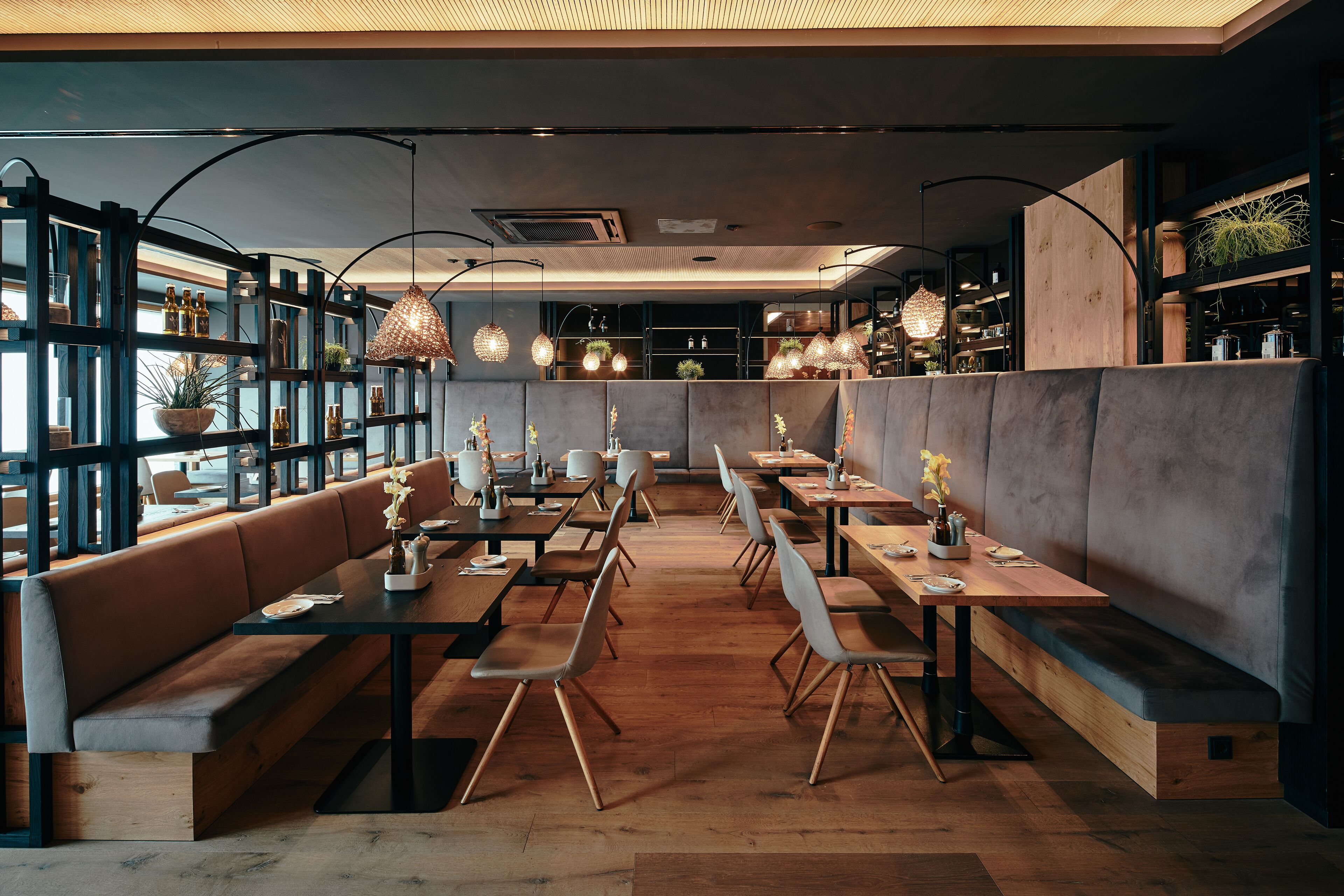 A modern restaurant interior with wooden tables, cushioned benches, and hanging pendant lights.