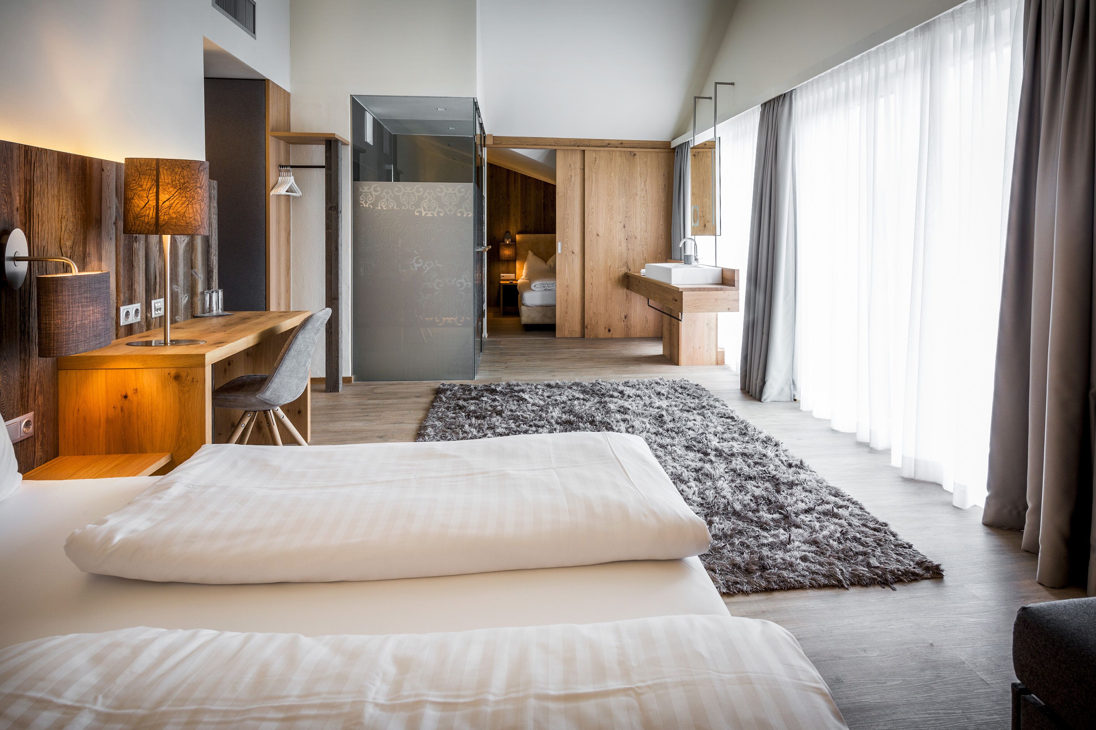 A modern hotel room with a large bed, a desk, a chair, a bathroom with a sink, and a large window with curtains.