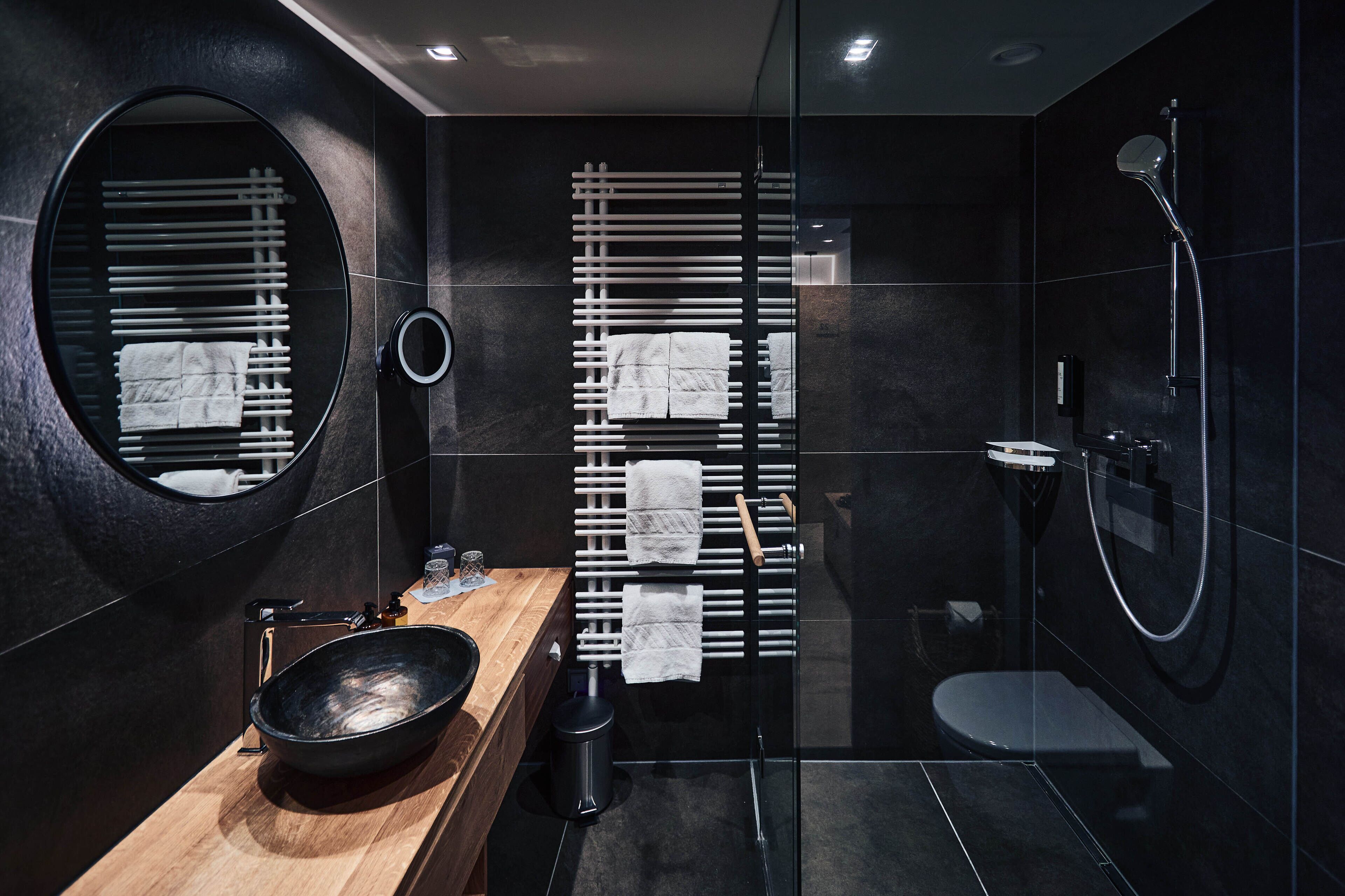 Un baño moderno con un espejo redondo, un mueble de tocador de madera, un lavabo negro, una mampara de ducha de vidrio y un inodoro.