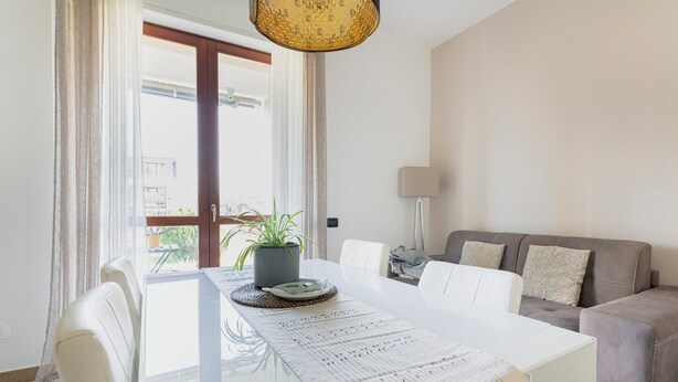 A modern living room with a glass dining table, white chairs, a grey sofa, and a potted plant. - HQ Aparthotel Milano Inn - Smart Suites (Cinisello Balsamo)