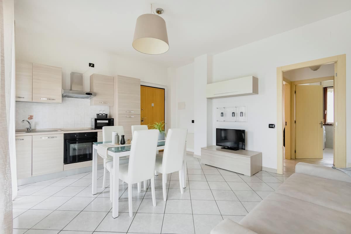 A modern kitchen with white cabinetry, a dining area with a glass table, and a living area with a sofa and TV.