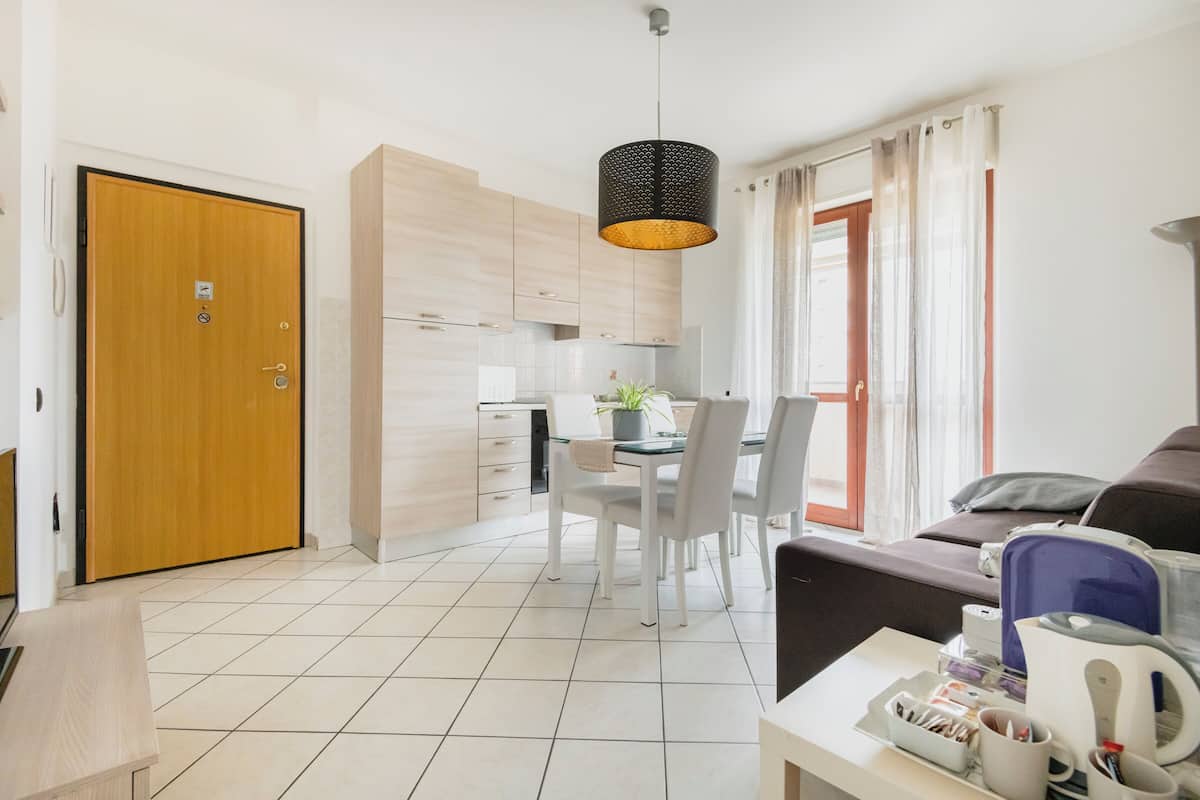 A modern kitchen with wooden cabinets, a dining table with chairs, a hanging pendant light, and a sofa in the living area.
