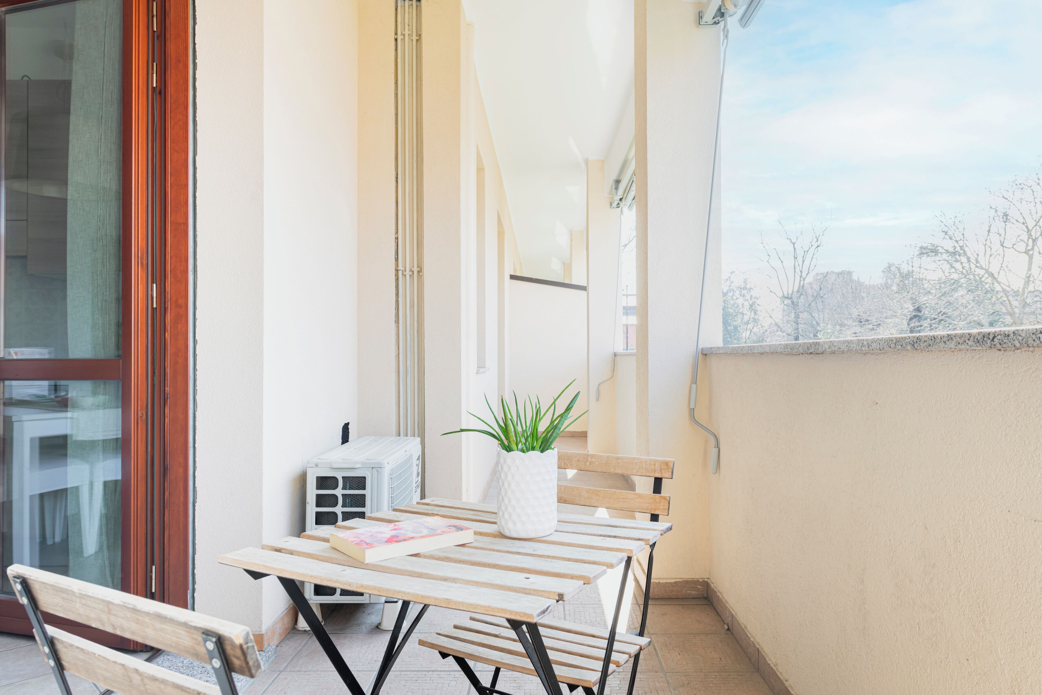 A balcony with a table, chairs, a potted plant, and a window with a view of trees.
