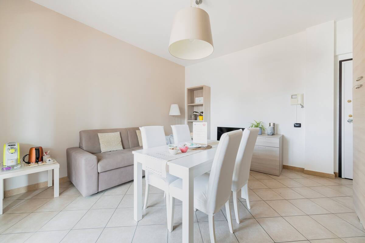 A modern, minimalist living room with a white sofa, a dining table with chairs, a coffee machine, and a bookshelf.