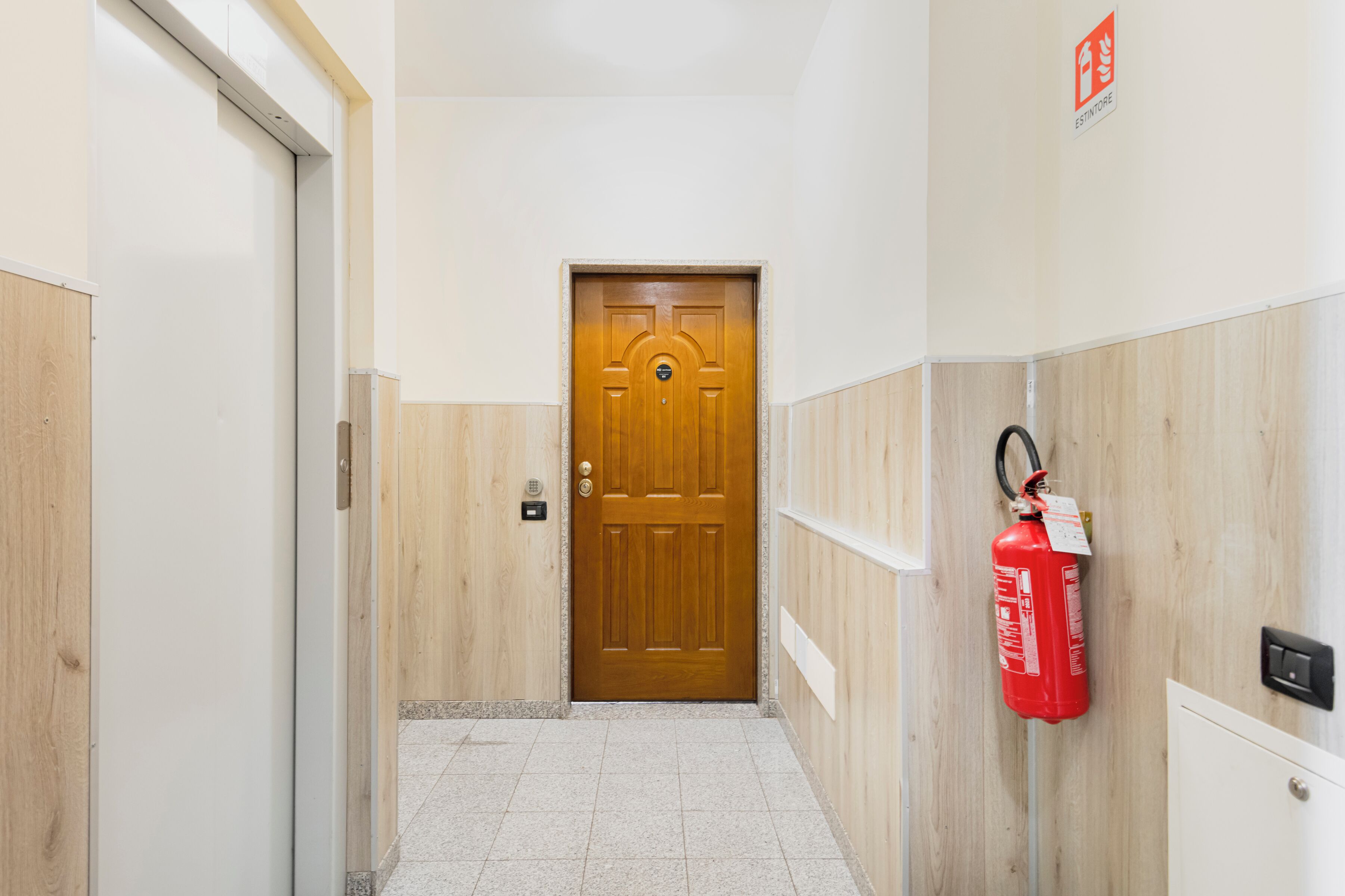 A hallway with a wooden door at the end, a fire extinguisher mounted on the wall, and a light switch.