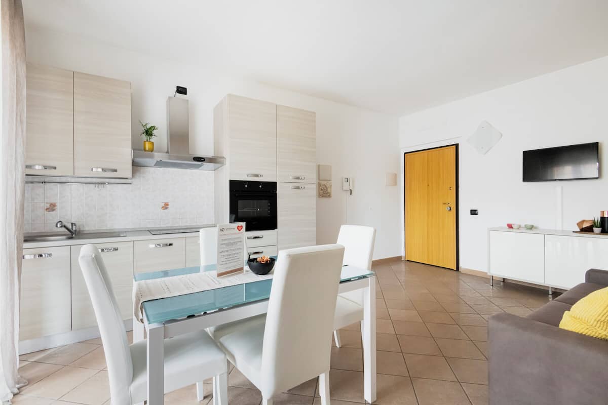 A modern kitchen with white cabinets, a dining table with chairs, a sofa, and a TV mounted on the wall.
