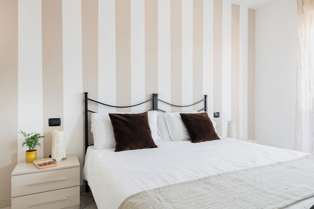 A neatly made bed with white linens and two brown pillows, a bedside table with a plant and a book, and a wall with vertical stripes.