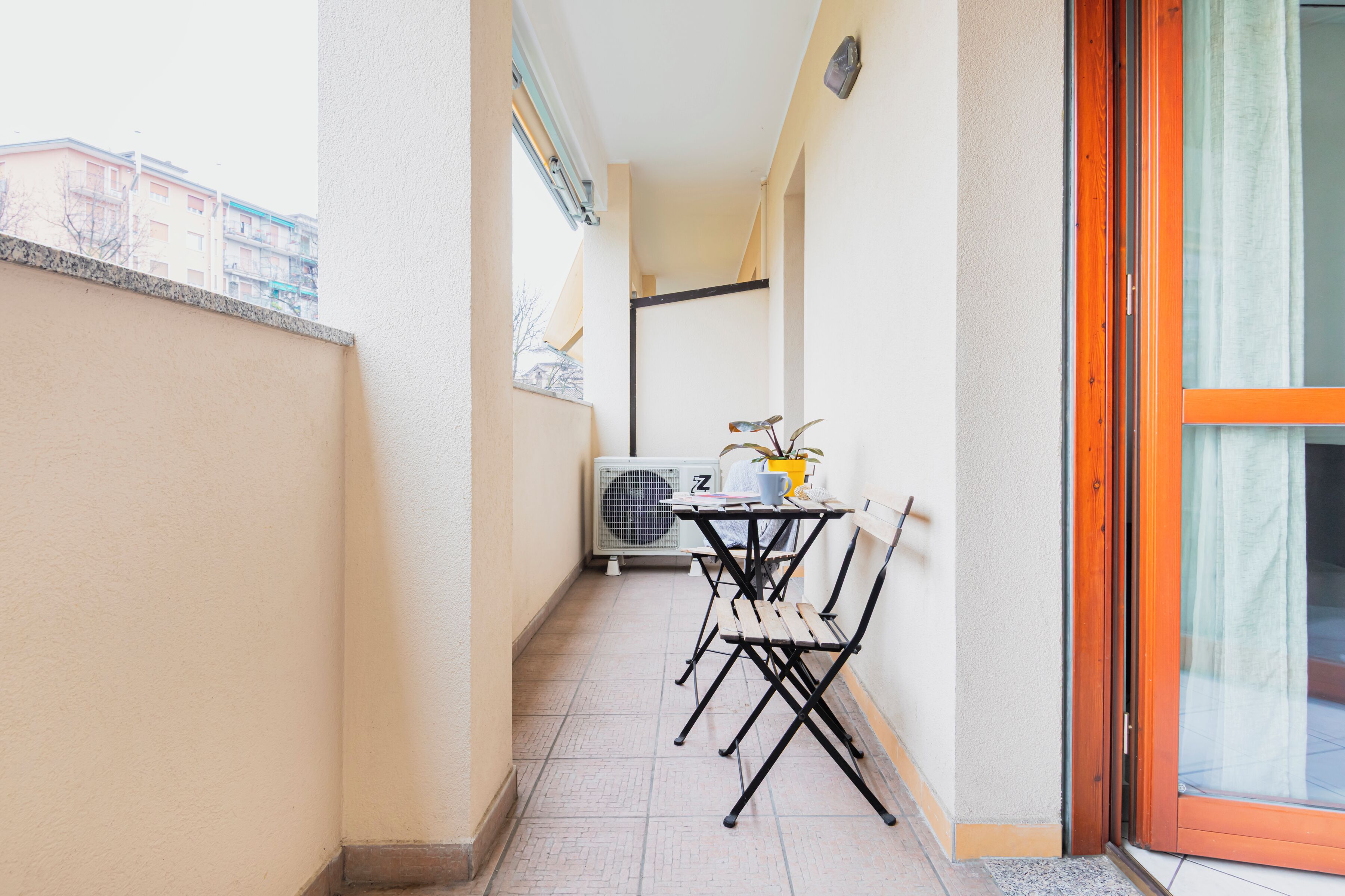 A narrow balcony with a table and chairs, an air conditioning unit, and a sliding glass door.