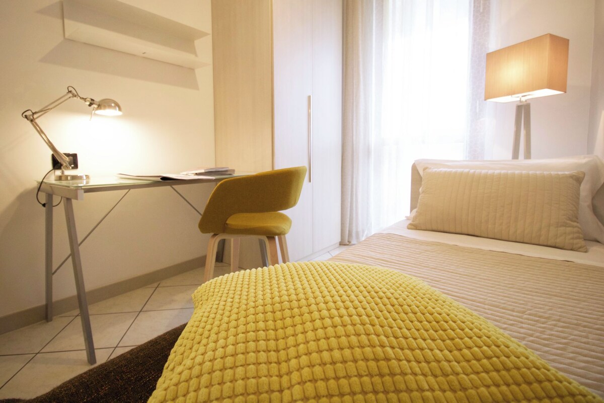A hotel room with a bed, a desk with a lamp, a chair, and a window with curtains.