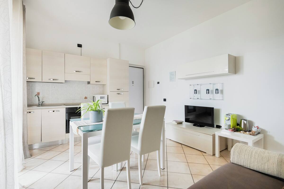 A modern kitchen with white cabinets, a dining table with chairs, a TV, and a small side table with a kettle and cups.