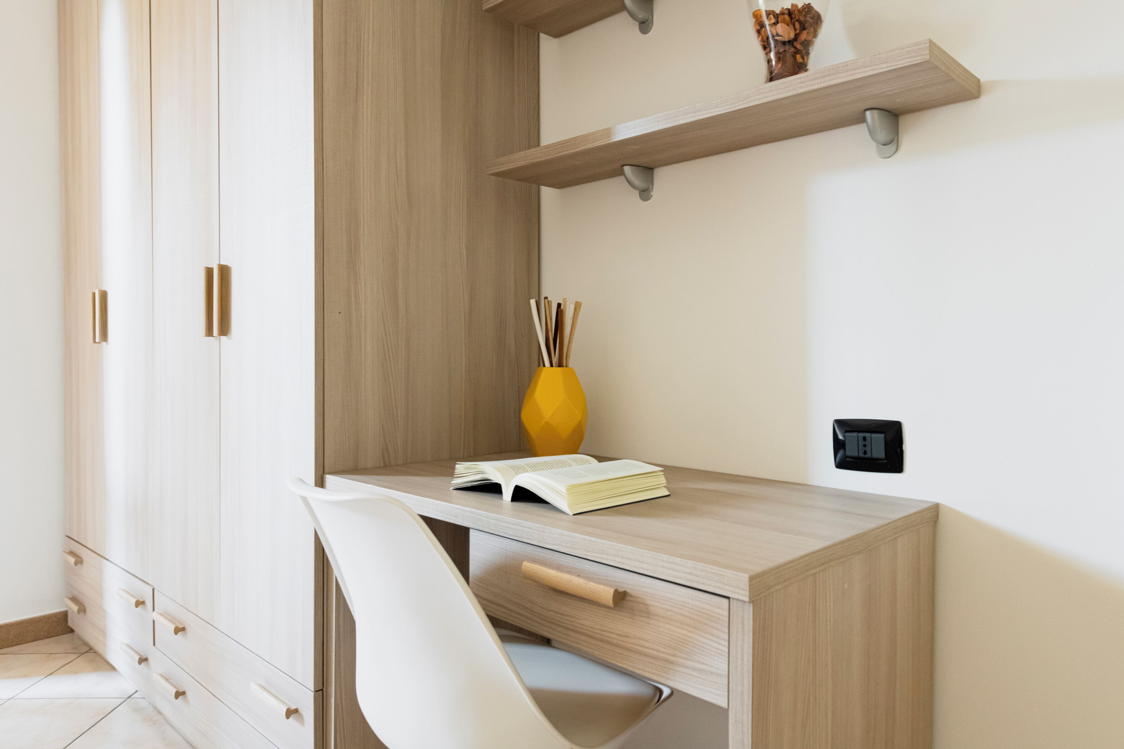 A modern study room with a wooden desk, a white chair, a book, a yellow vase, and a wall-mounted shelf.