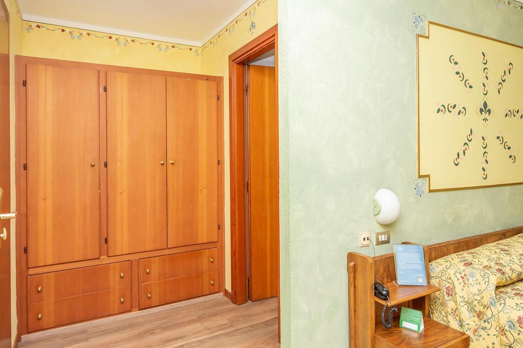 Ein Zimmer mit großem Holzschrank, einem Bett mit floraler Bettwäsche und einer Wandlampe.
