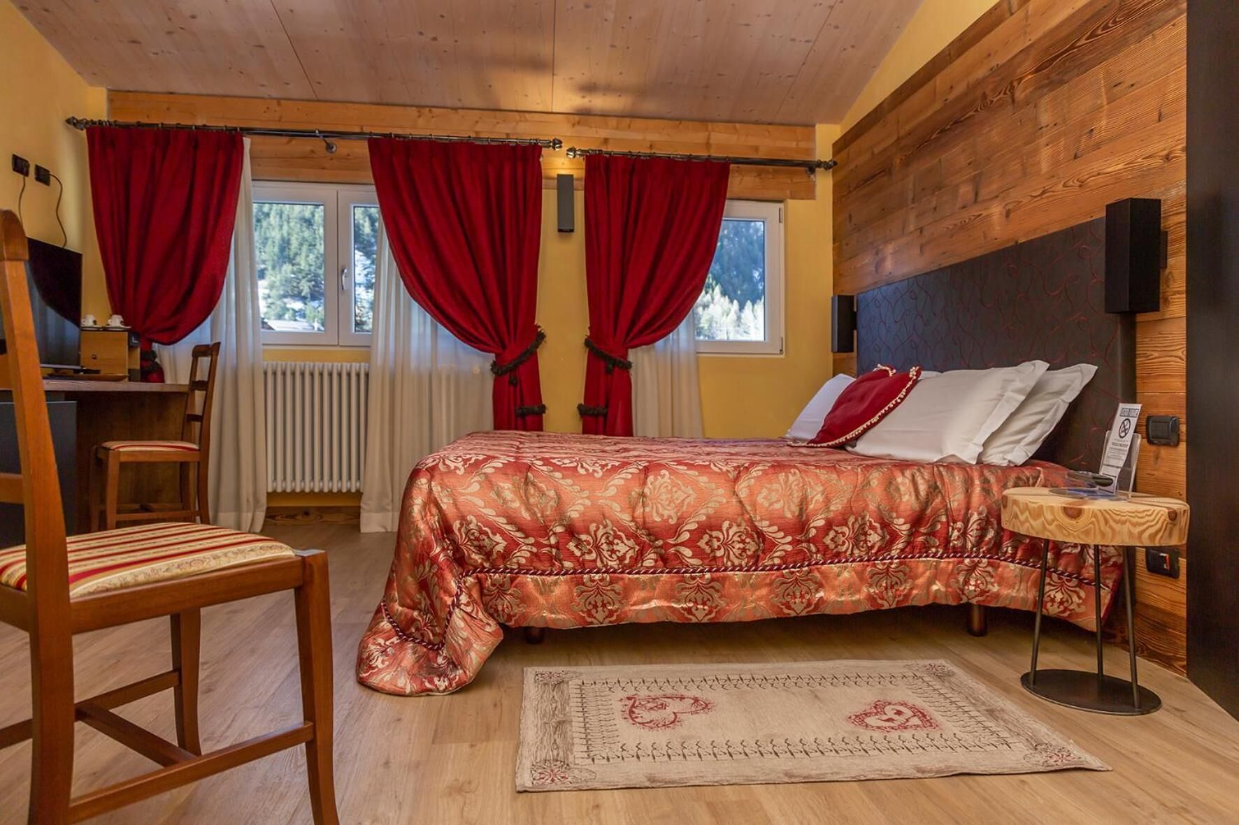 A cozy bedroom with a wooden headboard, a large bed with a floral bedspread, a wooden chair, a small table with a laptop, and a view of the outdoors through a window with red curtains.