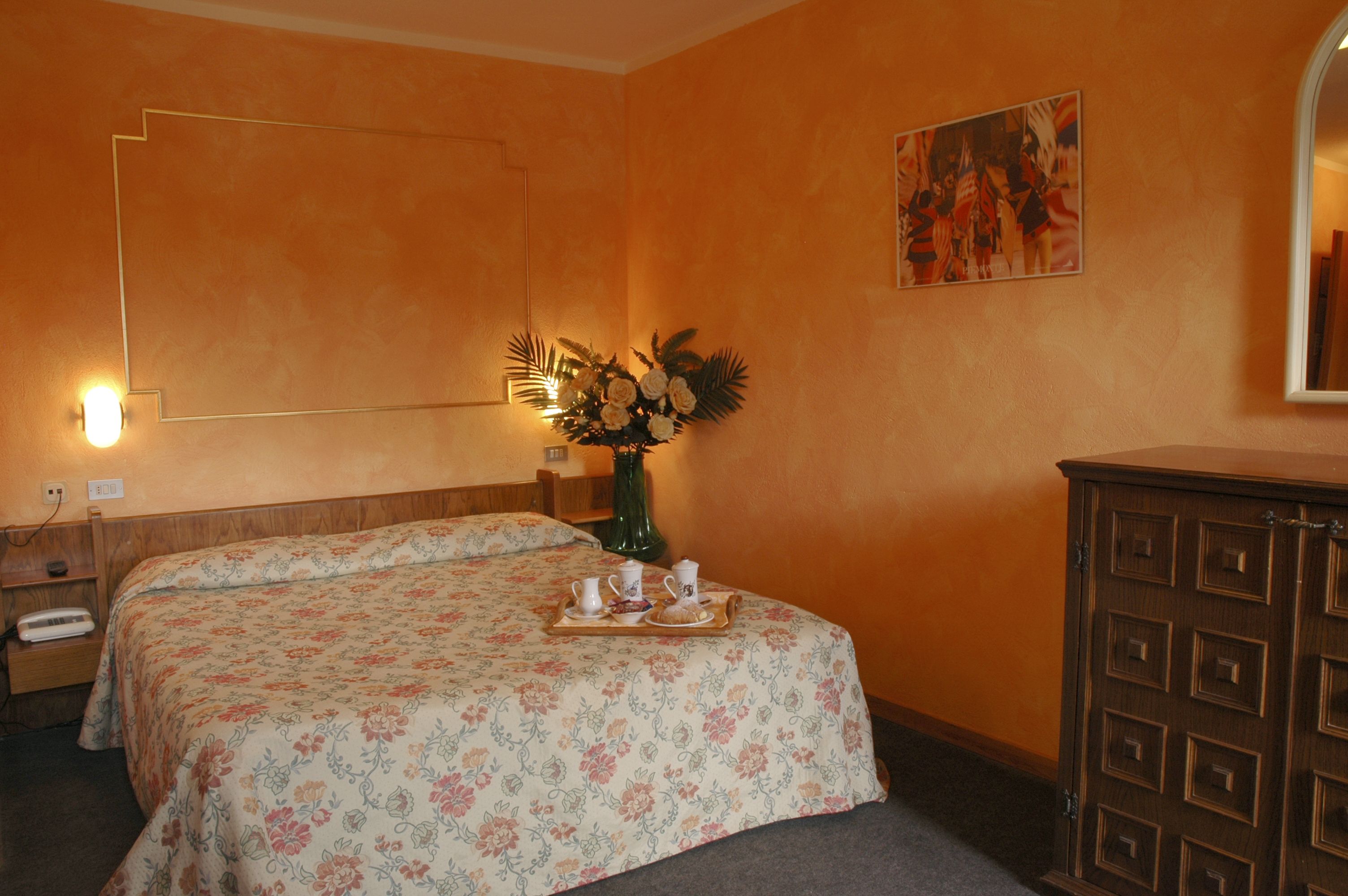 Habitación de hotel con colcha estampada con motivos florales, cómoda de madera, florero con flores y un cuadro en la pared.