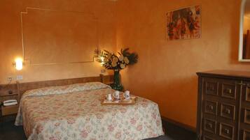 Habitación de hotel con colcha estampada con motivos florales, cómoda de madera, florero con flores y un cuadro en la pared.