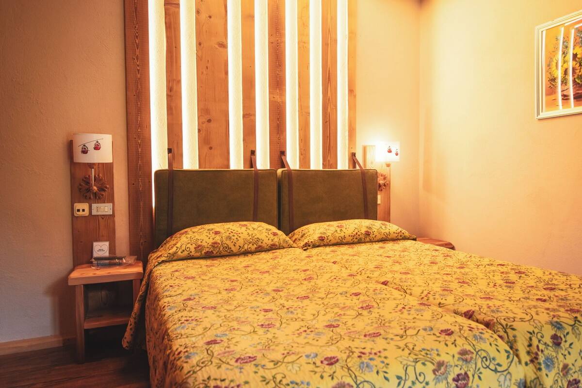 A bedroom with a wooden headboard, a floral bedspread, a nightstand with a lamp, and a wall-mounted light fixture.