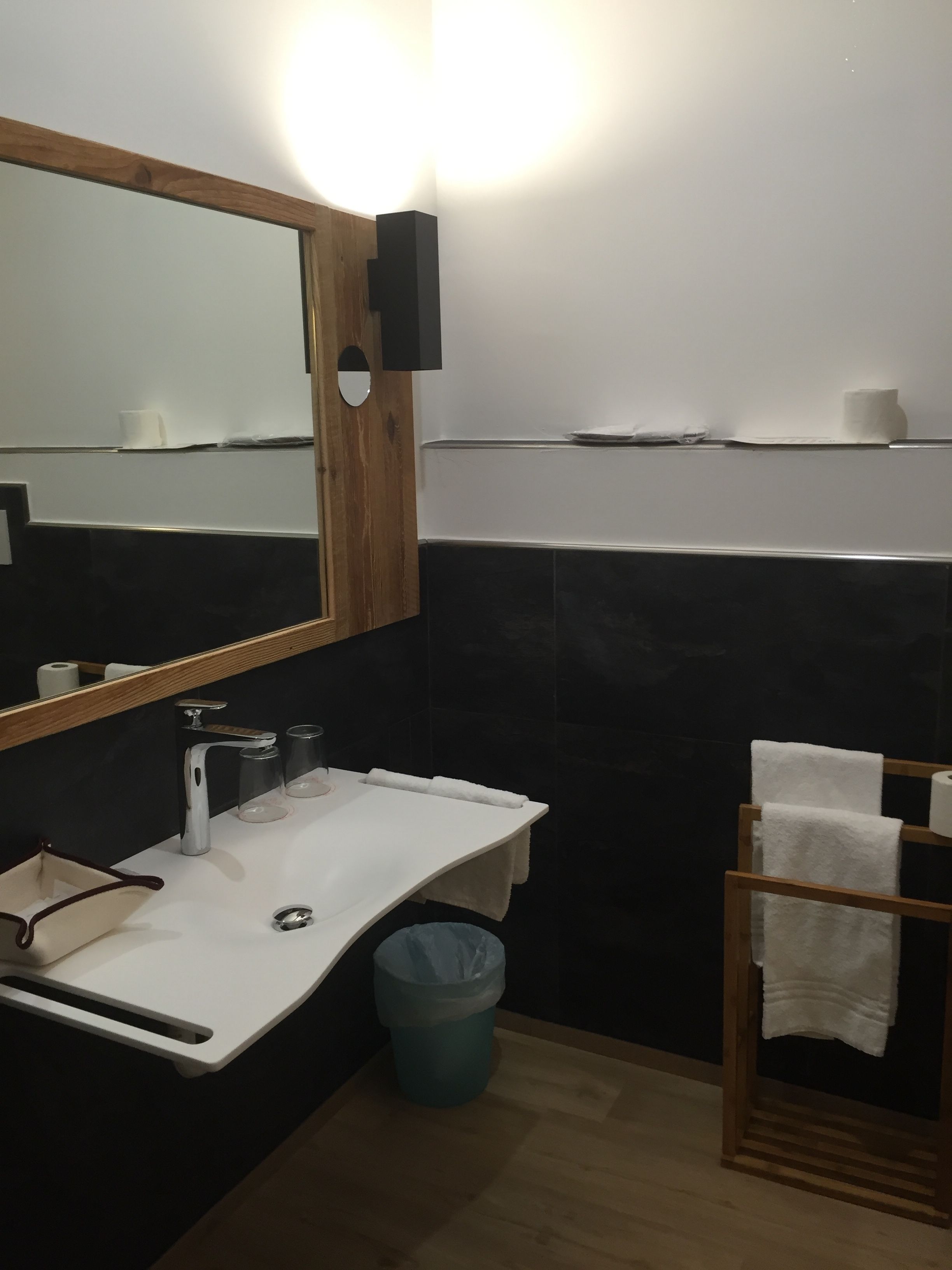 Un baño moderno con un lavamanos blanco, un espejo con marco de madera, un perchero para toallas y un cesto de basura.