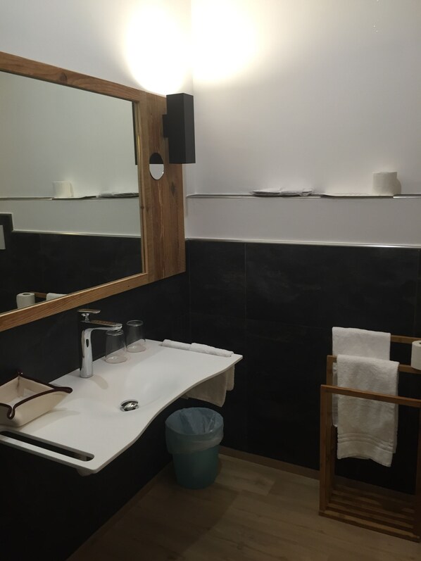Un baño moderno con un lavamanos blanco, un espejo con marco de madera, un perchero para toallas y un cesto de basura.